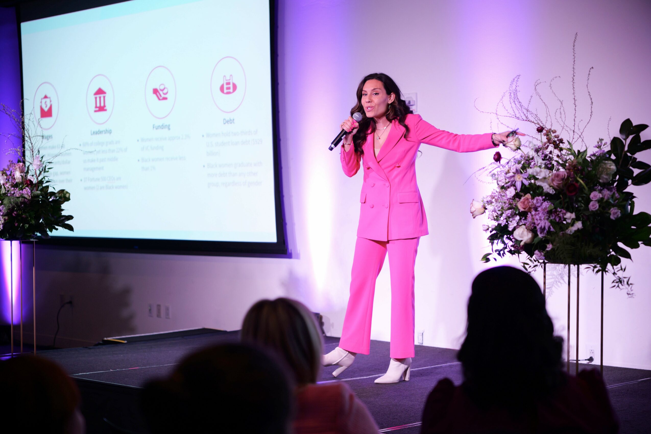 Woman in pink suit speaking on stage with flowers and presentation screen, audience visible in foreground.