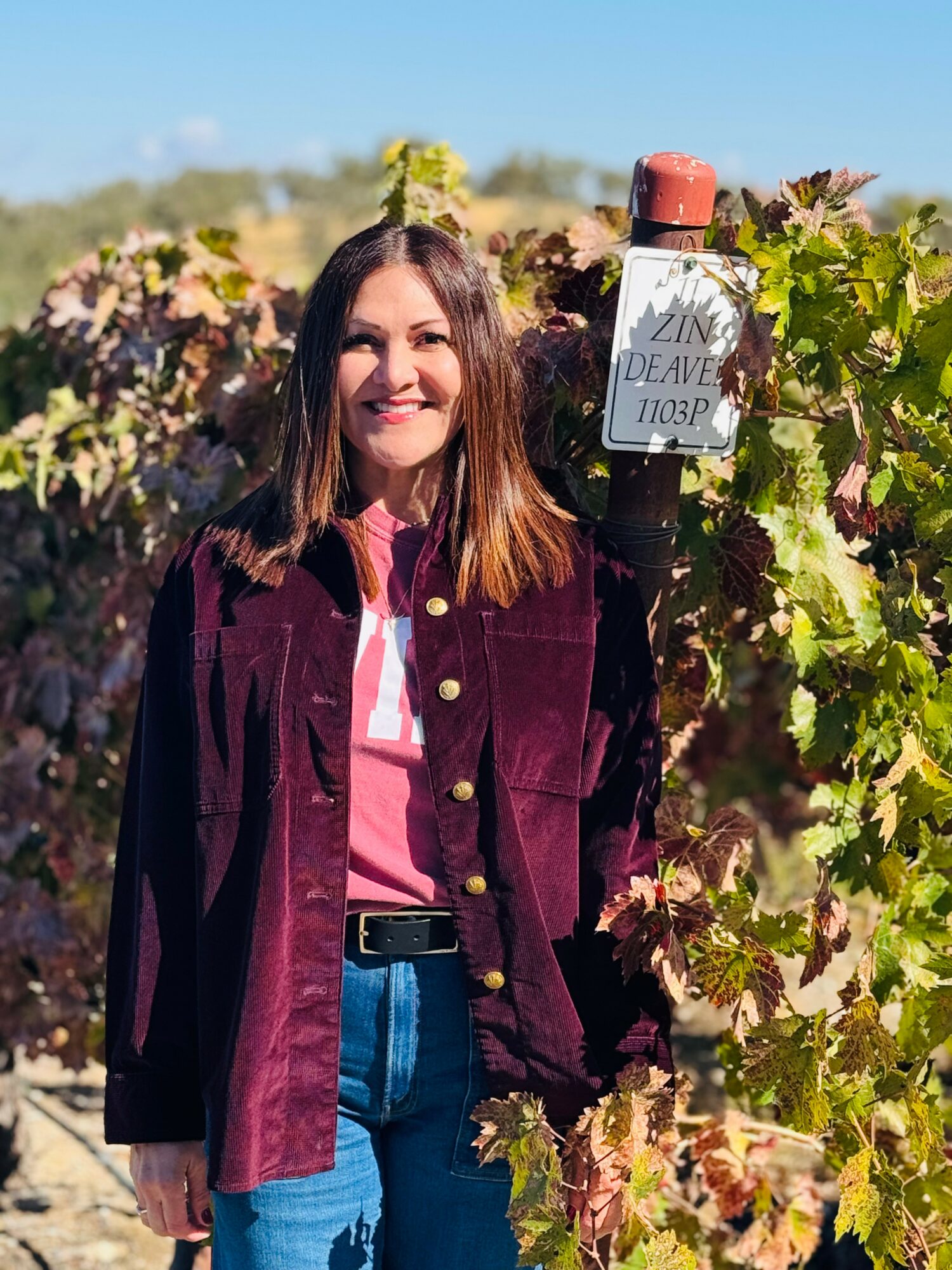 Woman smiling outdoors among grapevines, wearing a pink shirt and dark jacket, with a sign on a vine post.