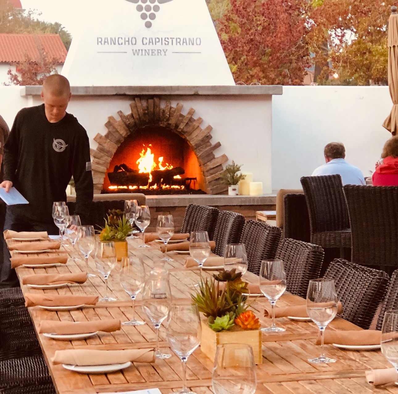 Outdoor dining table set with glasses, plates, and plants, with a pizza oven and a person in the background.