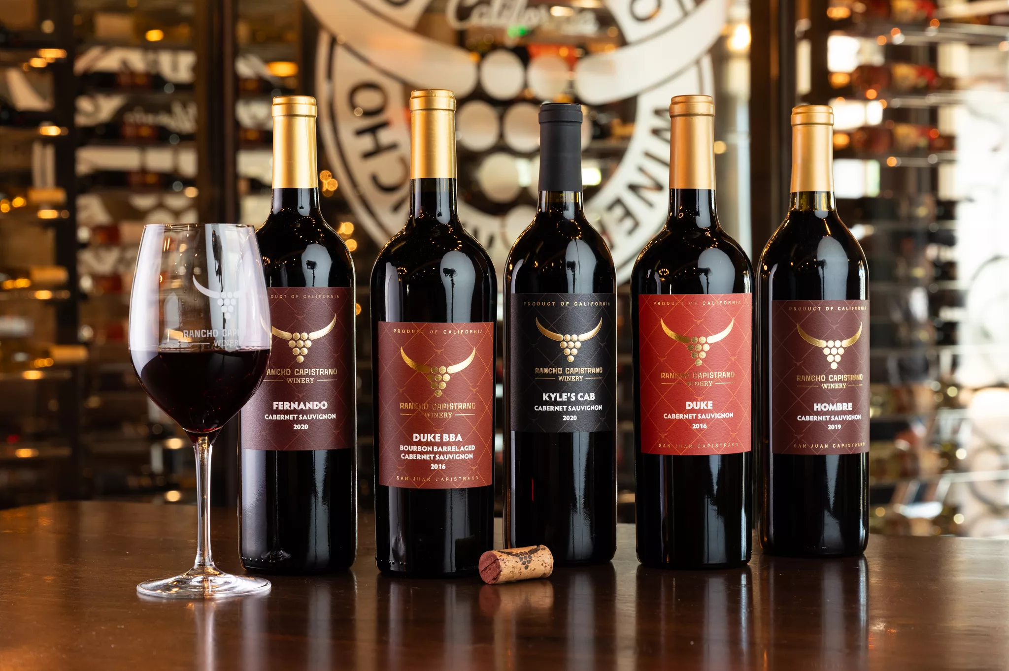 Five bottles of red wine and a glass of wine on a table, with a wine cellar background.