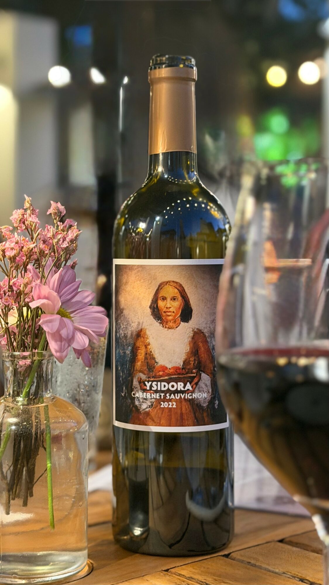A wine bottle with a label featuring a woman, placed on a table with flowers and a glass of red wine nearby.