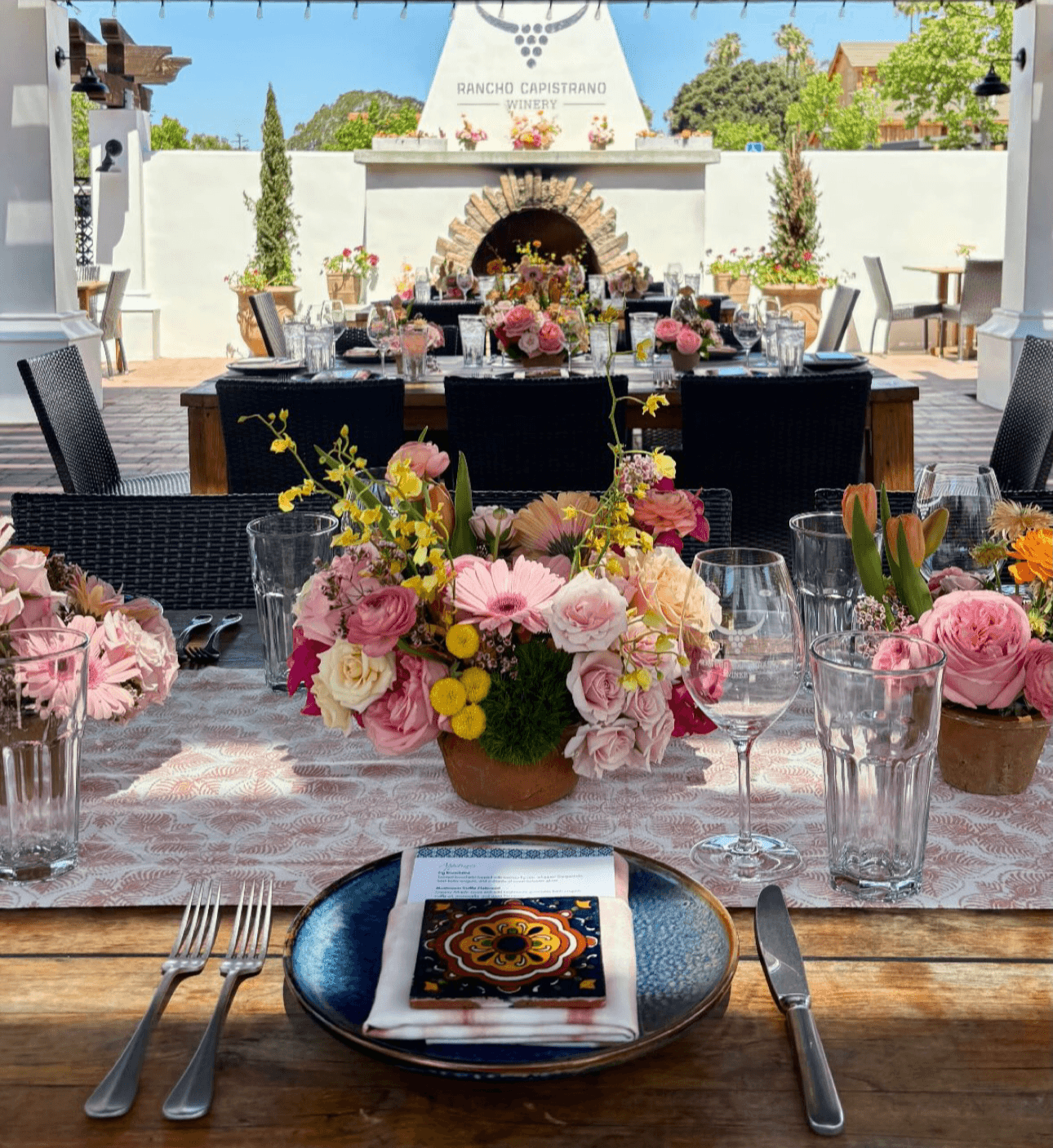 Elegant outdoor dining setup with floral centerpieces, glassware, and a fireplace in the background, under a clear sky.