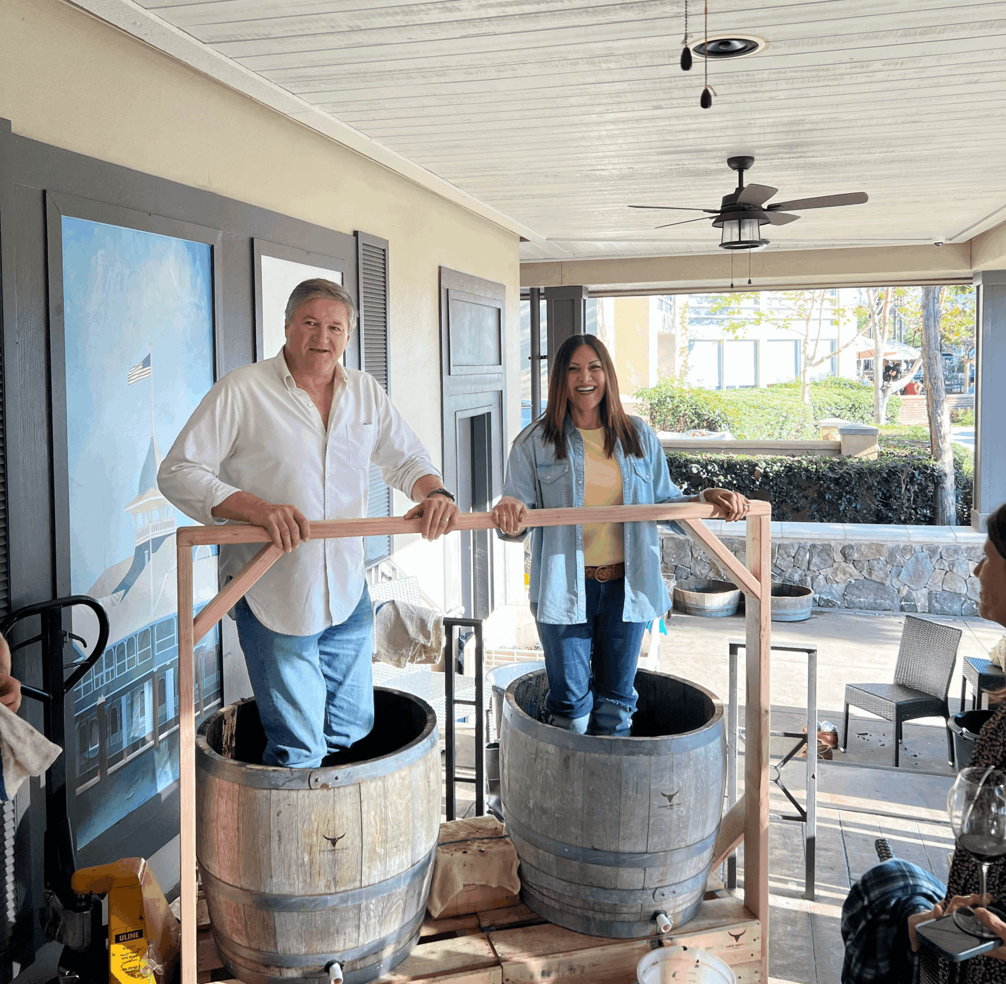 Two people stand inside large wooden barrels, holding a wooden bar, in a sunlit room with outdoor view.