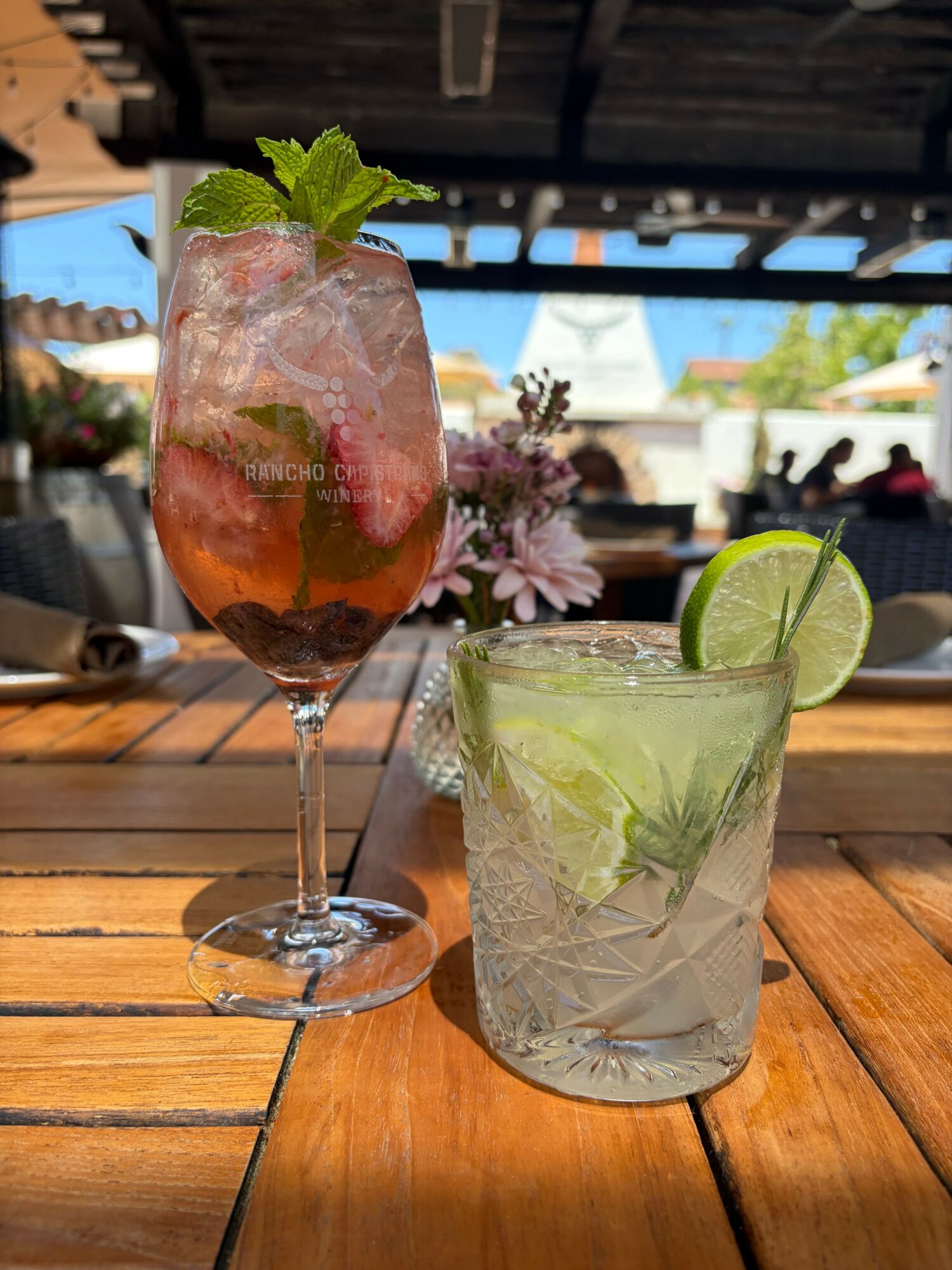 Two drinks on a wooden table, one with lime and mint, the other with a pink hue and mint, outdoors.
