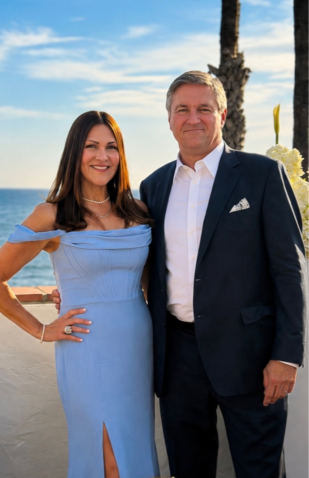 A woman in a light blue dress and a man in a dark suit stand outdoors near a beach with palm trees, smiling.