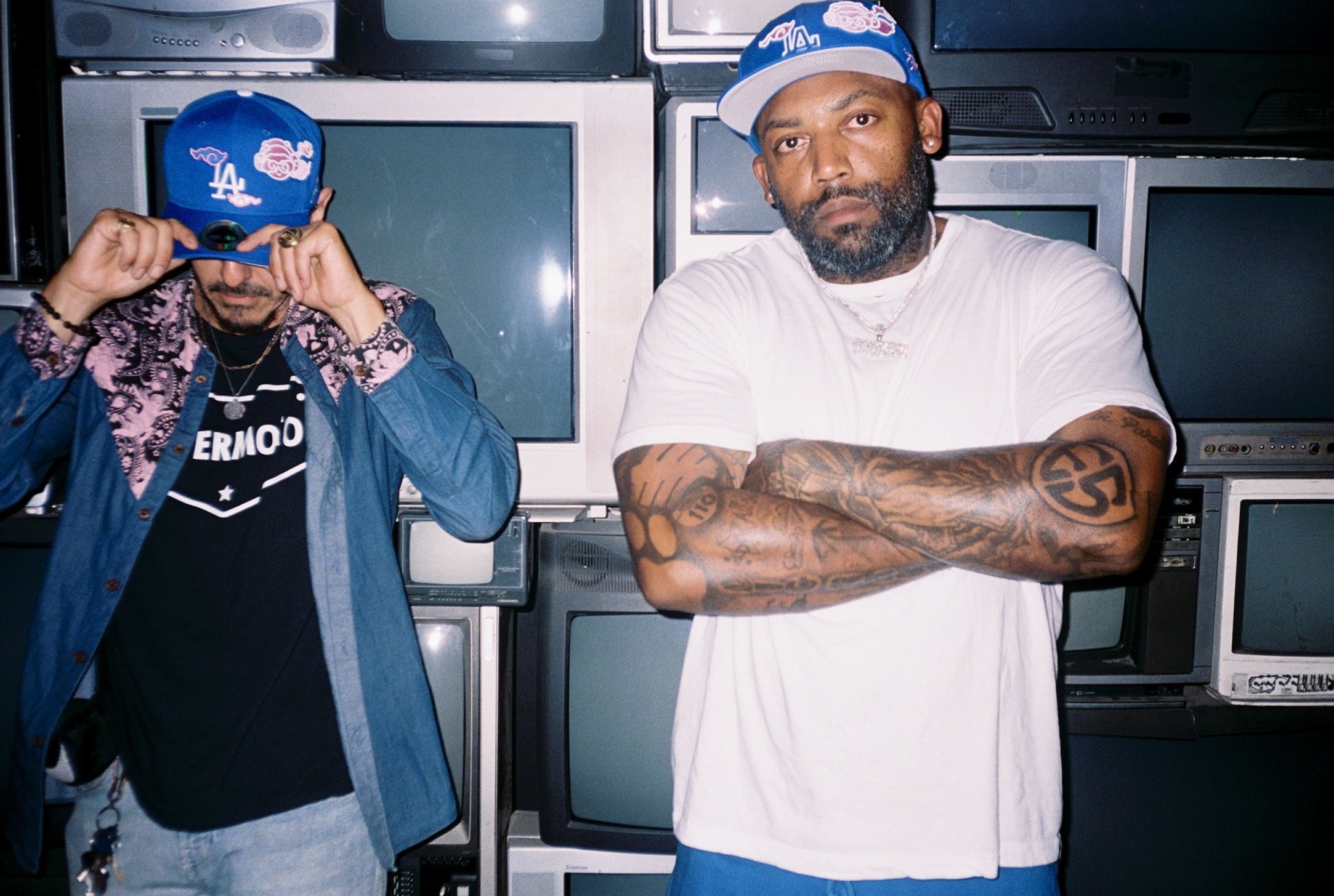Two men in a room with multiple old television screens; one wears a blue cap and white shirt, arms crossed, serious expression.