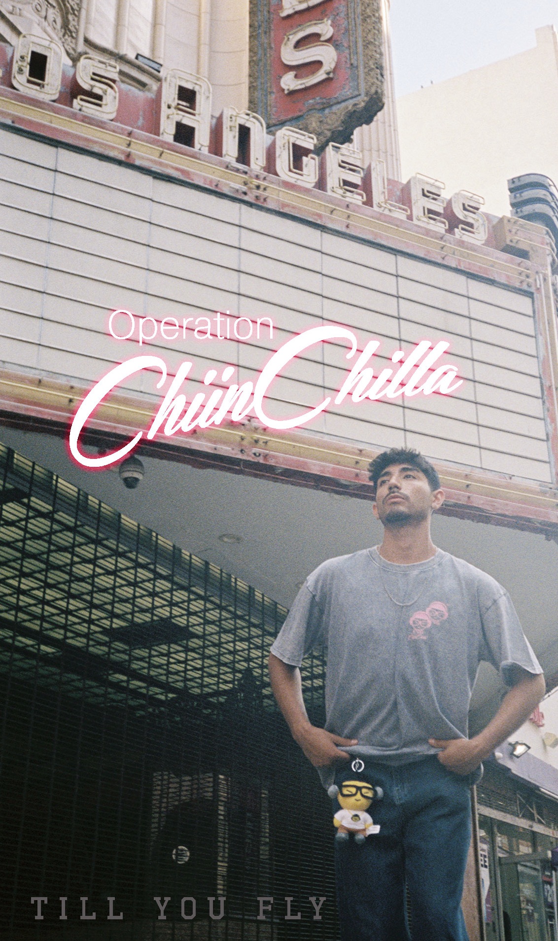 A man stands in front of a theater marquee with neon signs, wearing a gray T-shirt and holding a small plush toy.