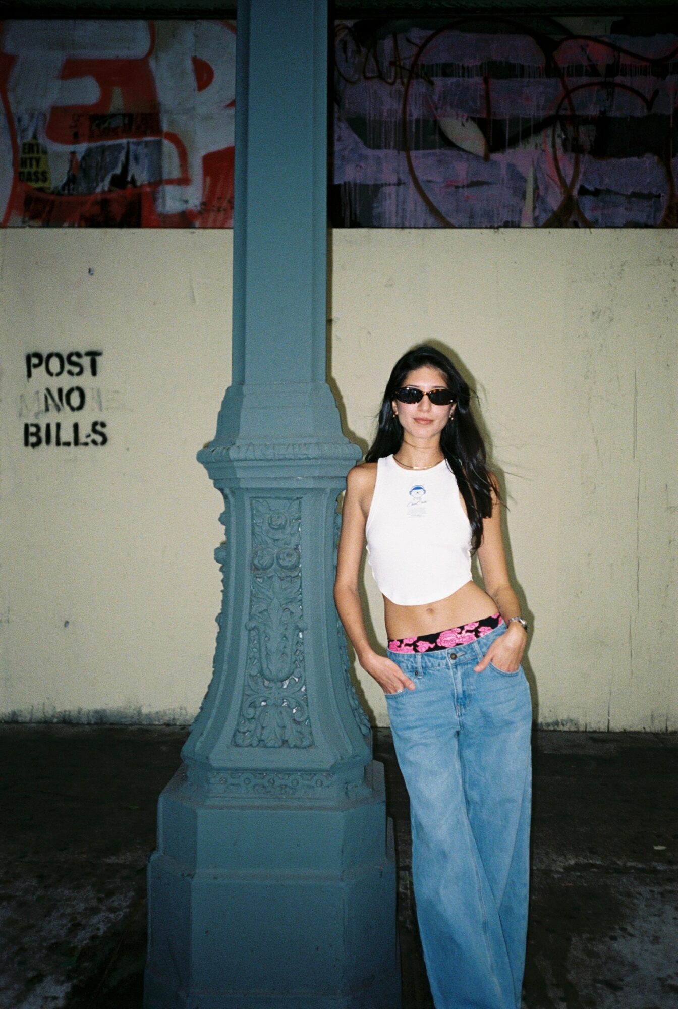 Young woman with long dark hair and sunglasses standing outdoors next to a blue lamppost, wearing a white top and blue jeans.