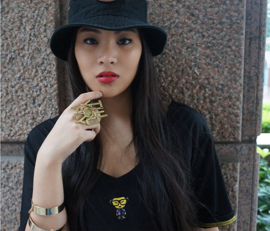 Young woman with long dark hair wearing a black hat and black shirt, standing against a stone wall.
