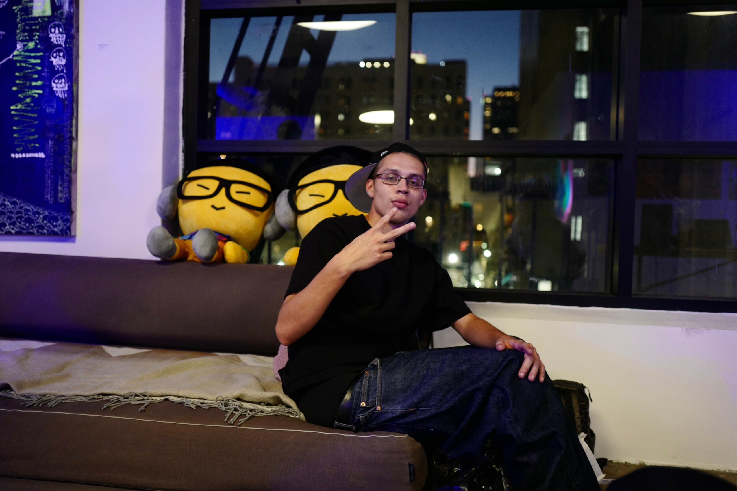 Person sitting on a bench with two plush toys behind, making a peace sign gesture, in a room with large windows and city lights outside.
