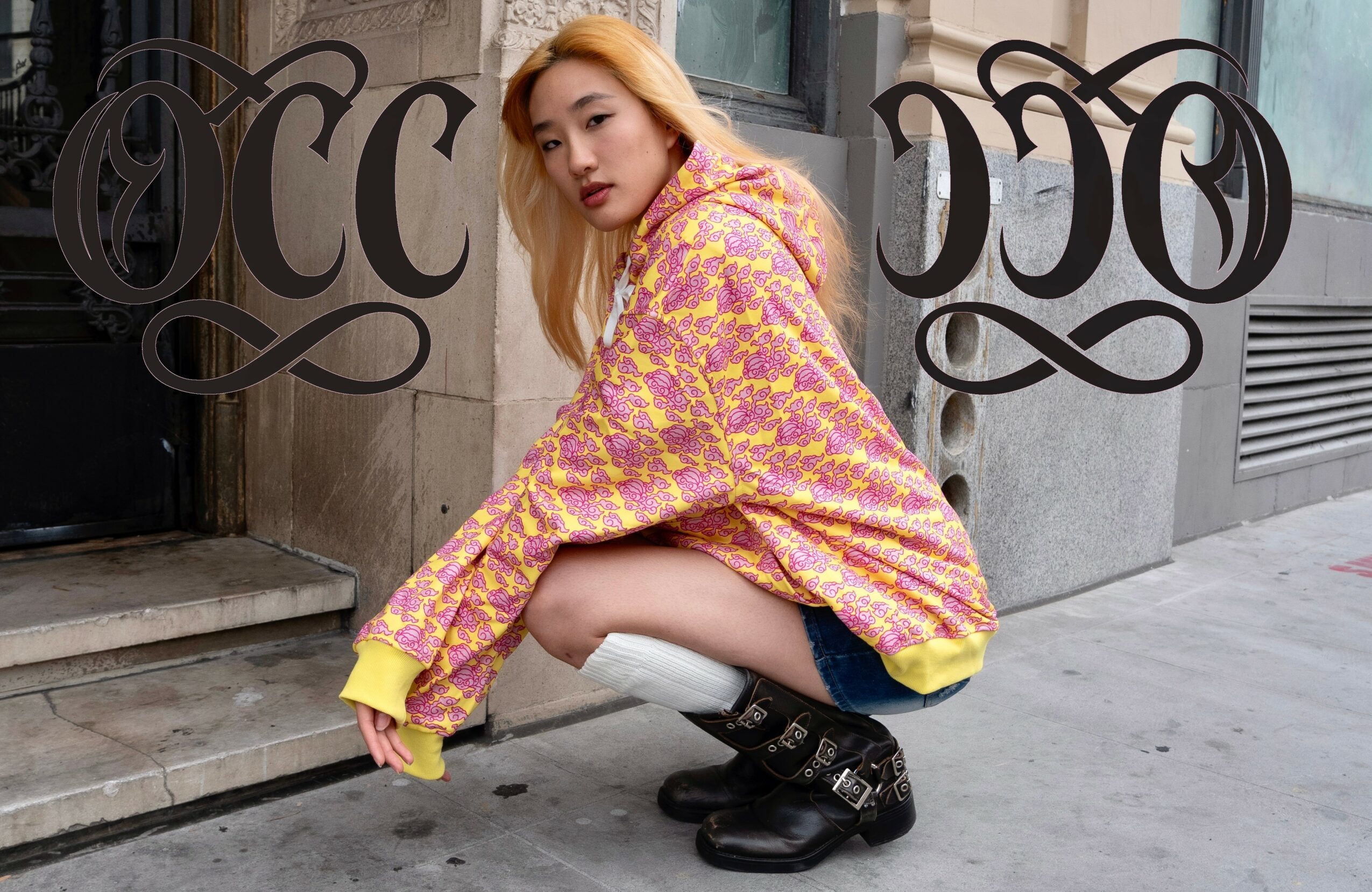Person crouching on sidewalk in colorful hoodie and black boots, urban background with building and stairs.