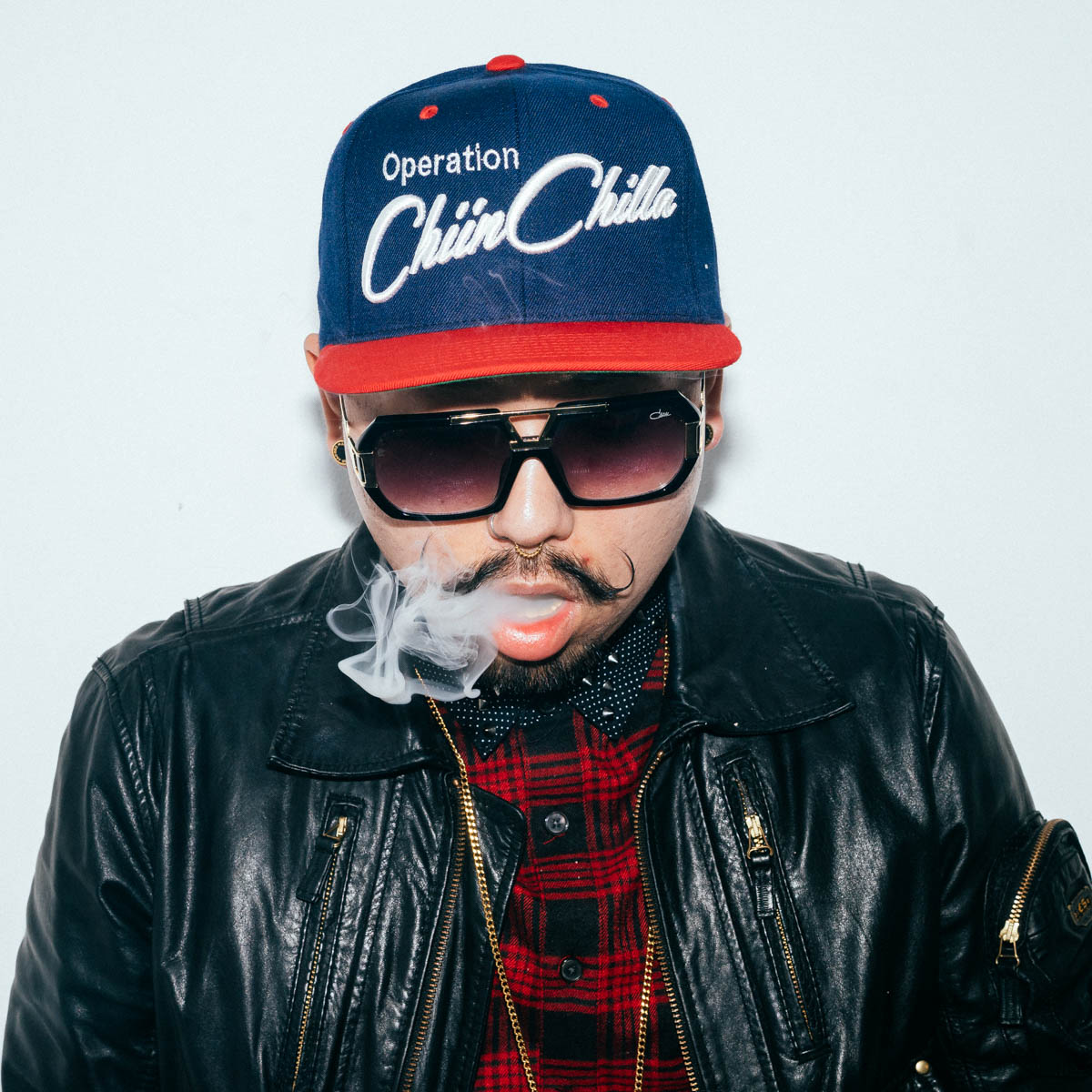 Person wearing sunglasses, a baseball cap, a leather jacket, and a red plaid shirt, smoking with smoke visible.