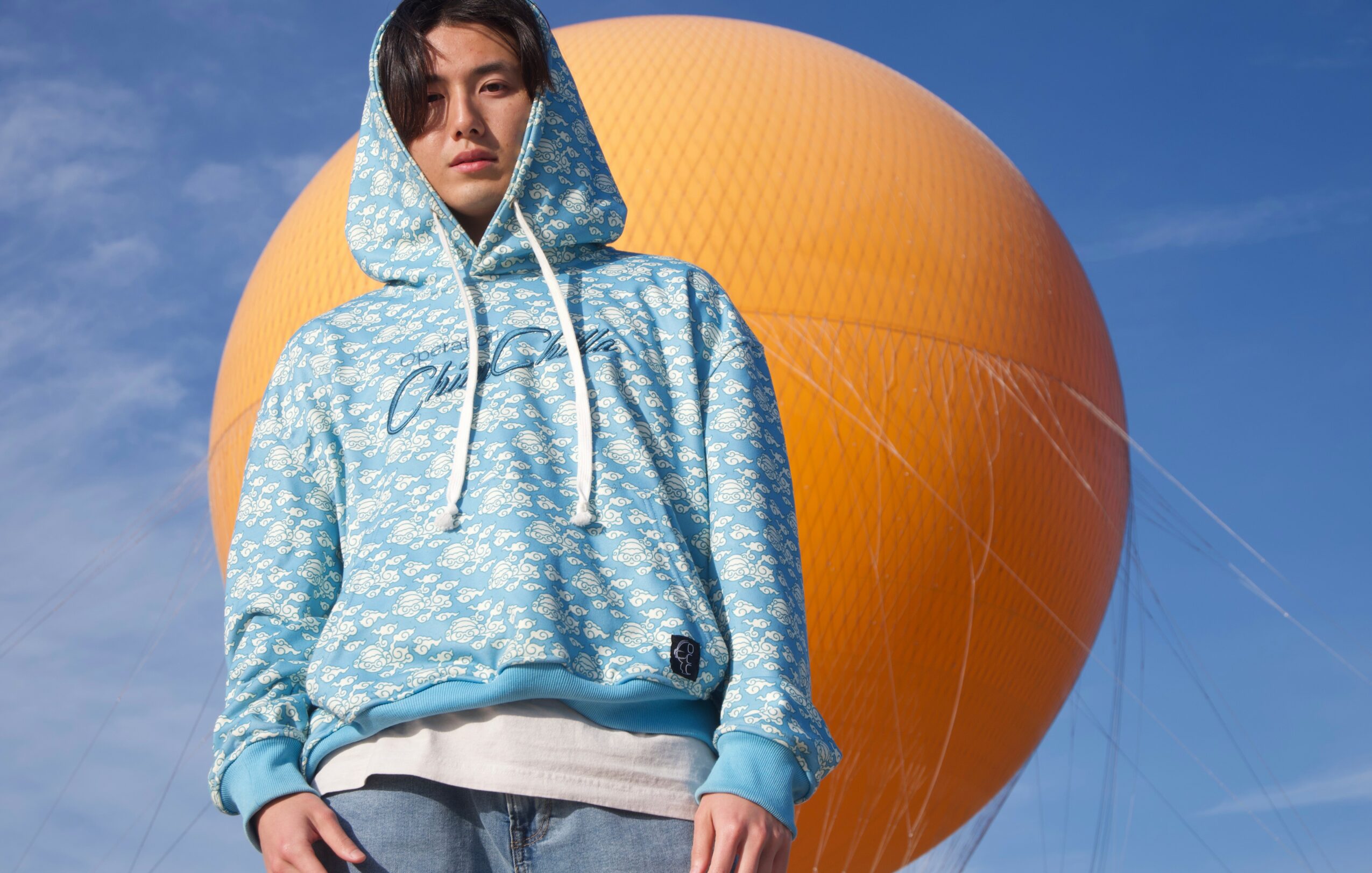 Young person in a blue patterned hoodie stands outdoors with a large orange hot air balloon behind them, blue sky above.