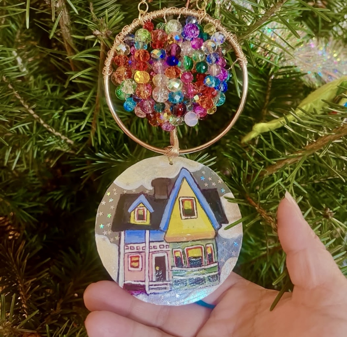 Hand holding a circular ornament with colorful beads and a painted house scene, hanging on a green tree.
