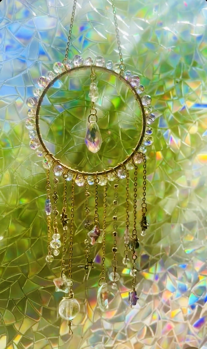 A circular dreamcatcher with hanging beads and a pink gemstone, set against a colorful, iridescent background.