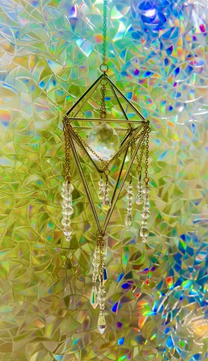 Hanging geometric ornament with beads and crystals against a colorful, holographic background.