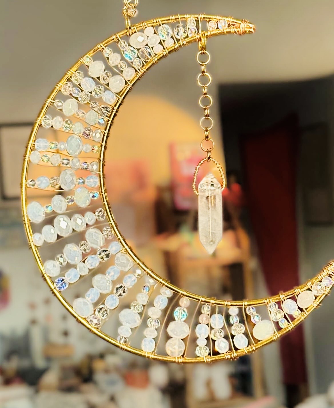 Decorative crescent moon ornament with hanging crystal, gold frame, and numerous small beads, hanging indoors with blurred background.