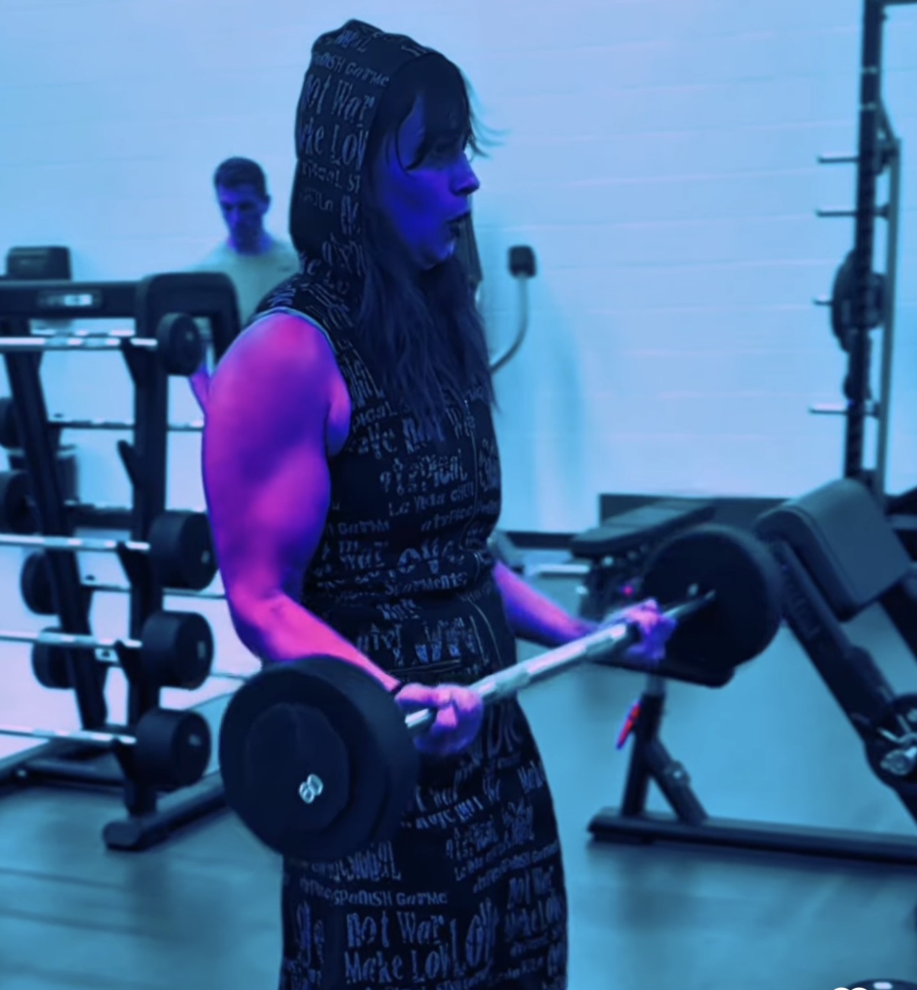 Woman lifting a dumbbell in a gym with blue lighting, wearing a sleeveless hoodie, with gym equipment in background.