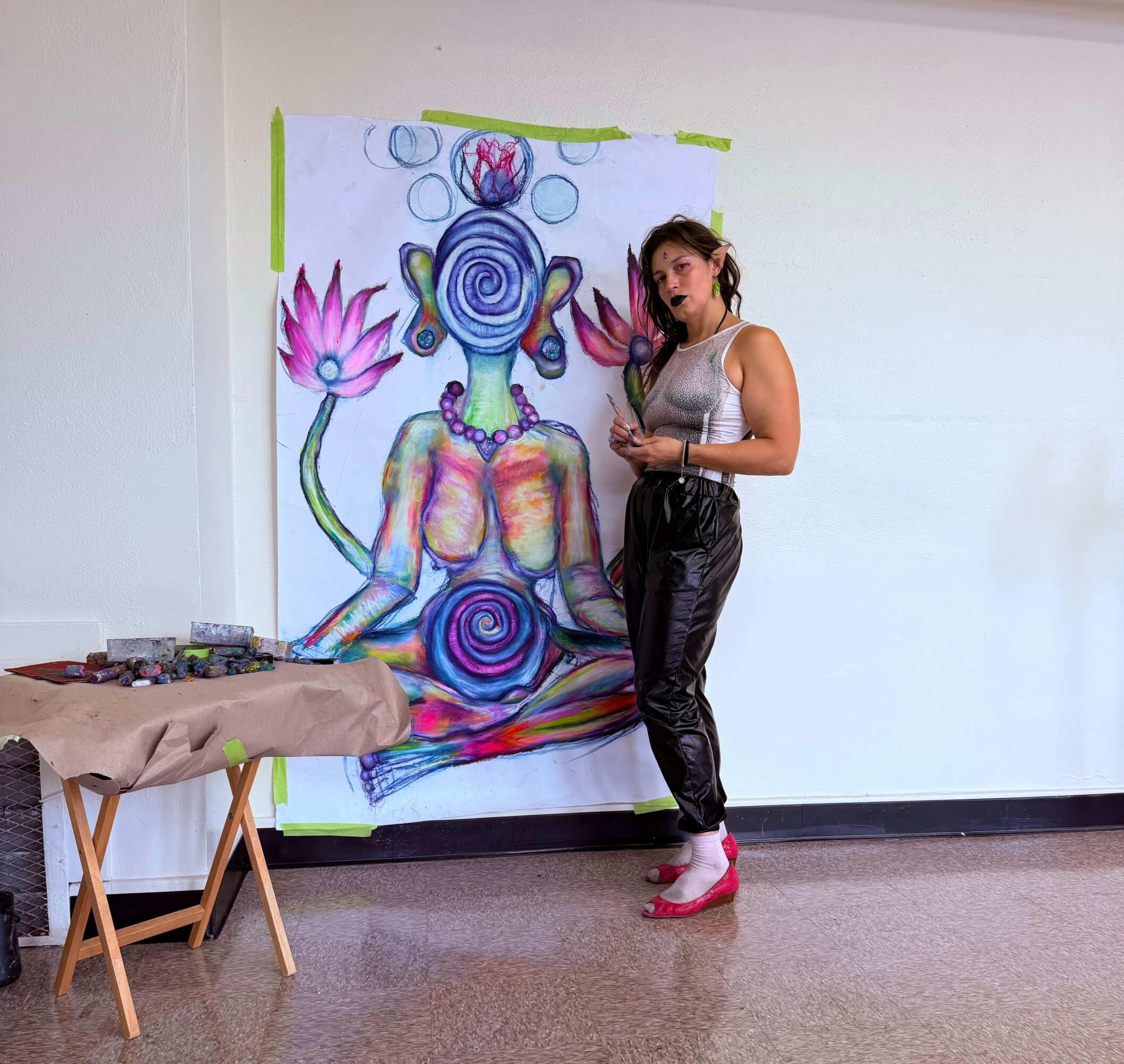 Woman standing next to colorful abstract painting on wall, holding a phone, with art supplies on table nearby.