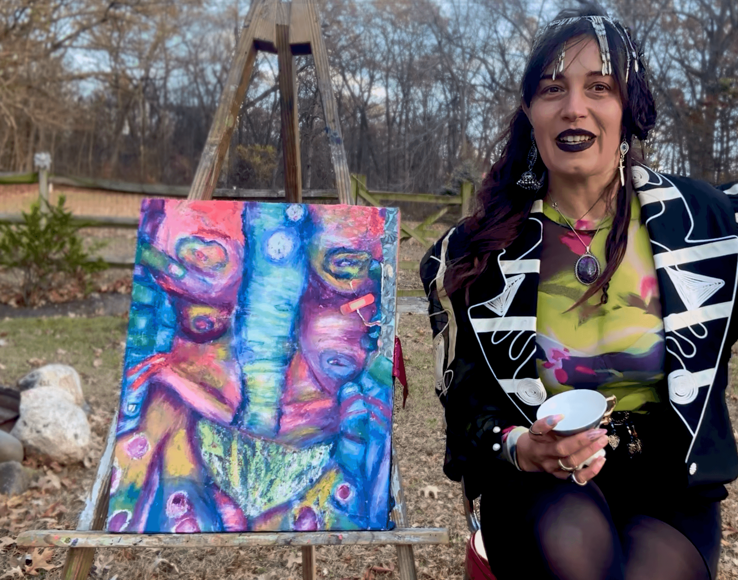 Woman with dark lipstick and earrings standing outdoors next to colorful abstract painting on easel, holding a cup.
