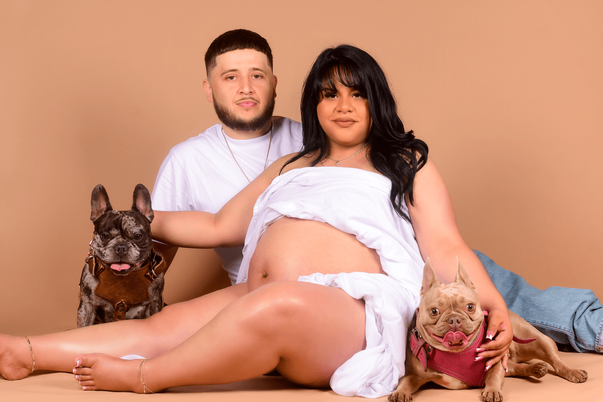 A pregnant woman with long dark hair and a man with short hair sit with two dogs against a beige background.