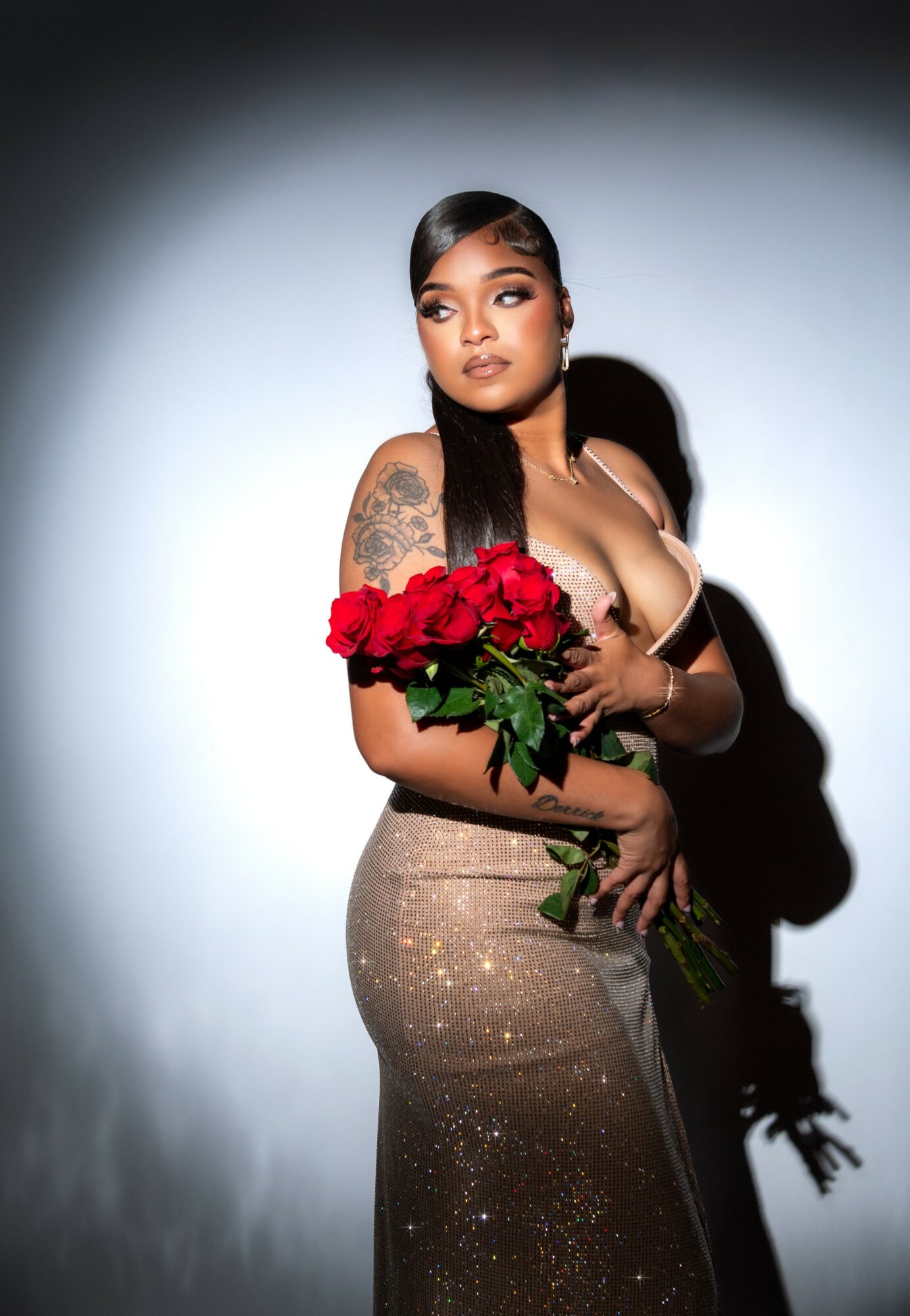 Woman with long dark hair holding a bouquet of red roses, wearing a sparkly gold dress, standing against a plain background.