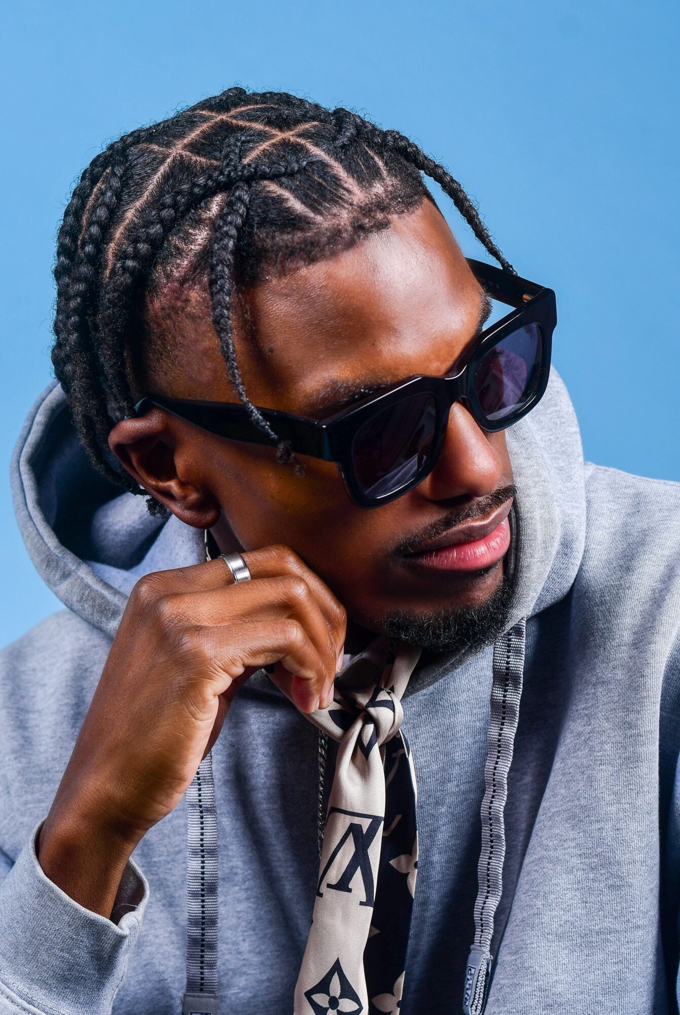 Man with braided hair wearing sunglasses and a gray hoodie, resting his chin on his hand against a blue background.