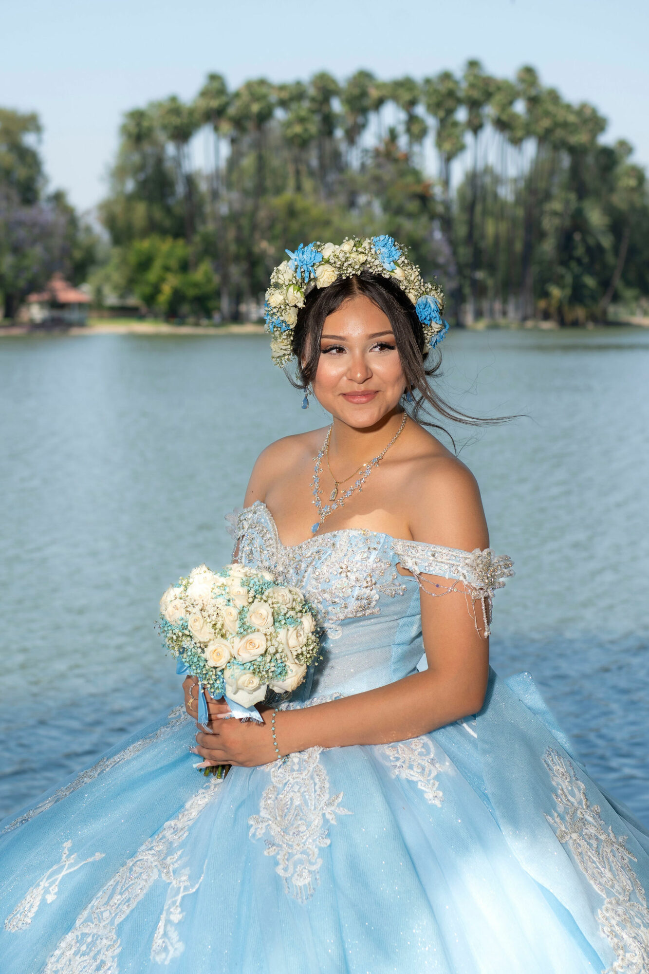 Young woman in a light blue dress with lace details, holding a bouquet of white flowers, standing outdoors near water with trees in background.
