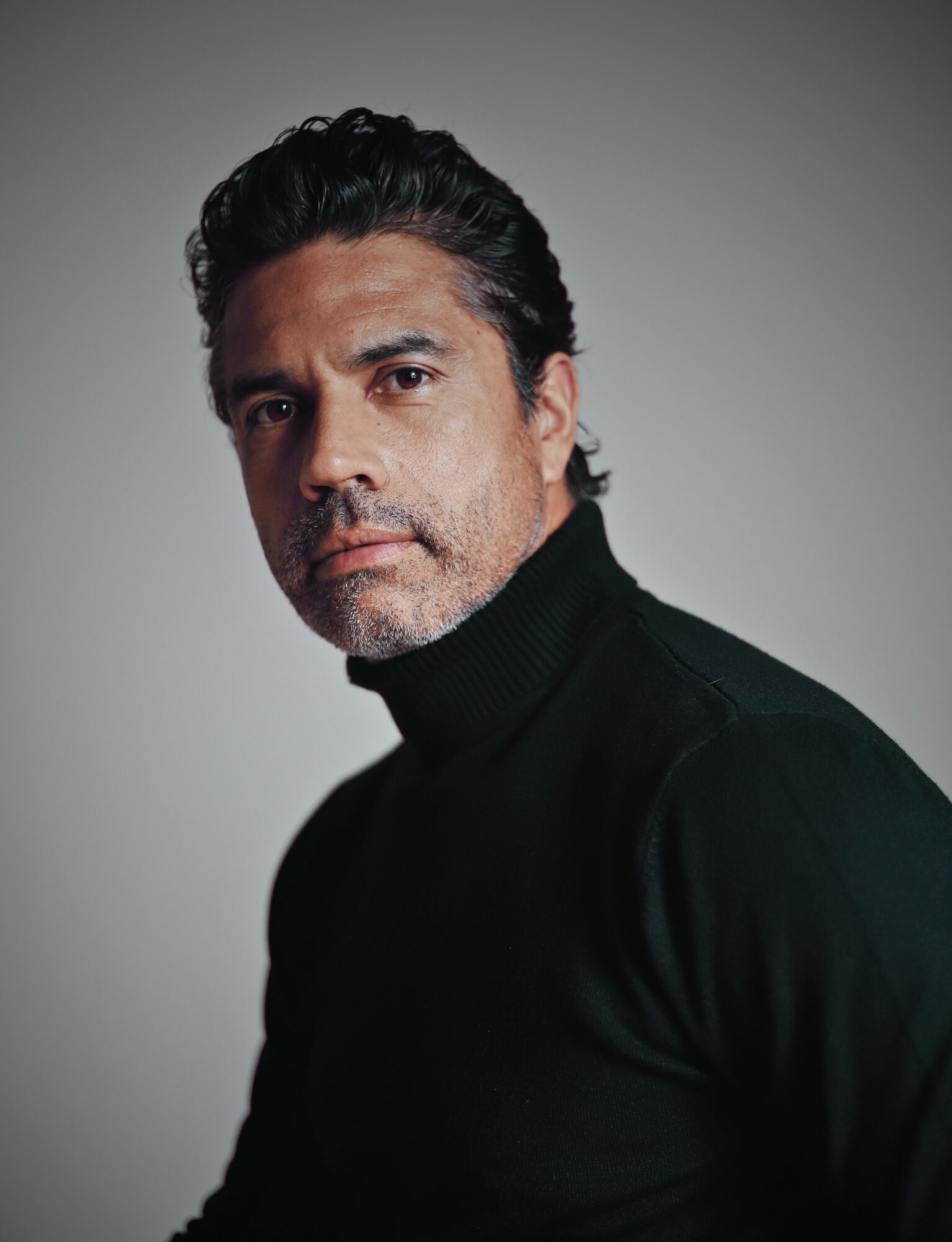 Portrait of a man with dark hair and stubble, wearing a black turtleneck, looking at the camera against a plain background.