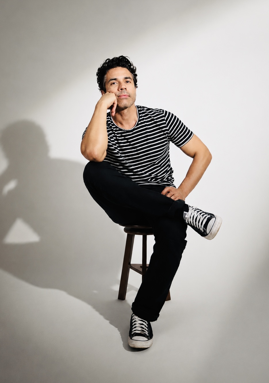 Young man sitting on a stool, resting his head on his hand, wearing a striped shirt and sneakers, against a plain background.