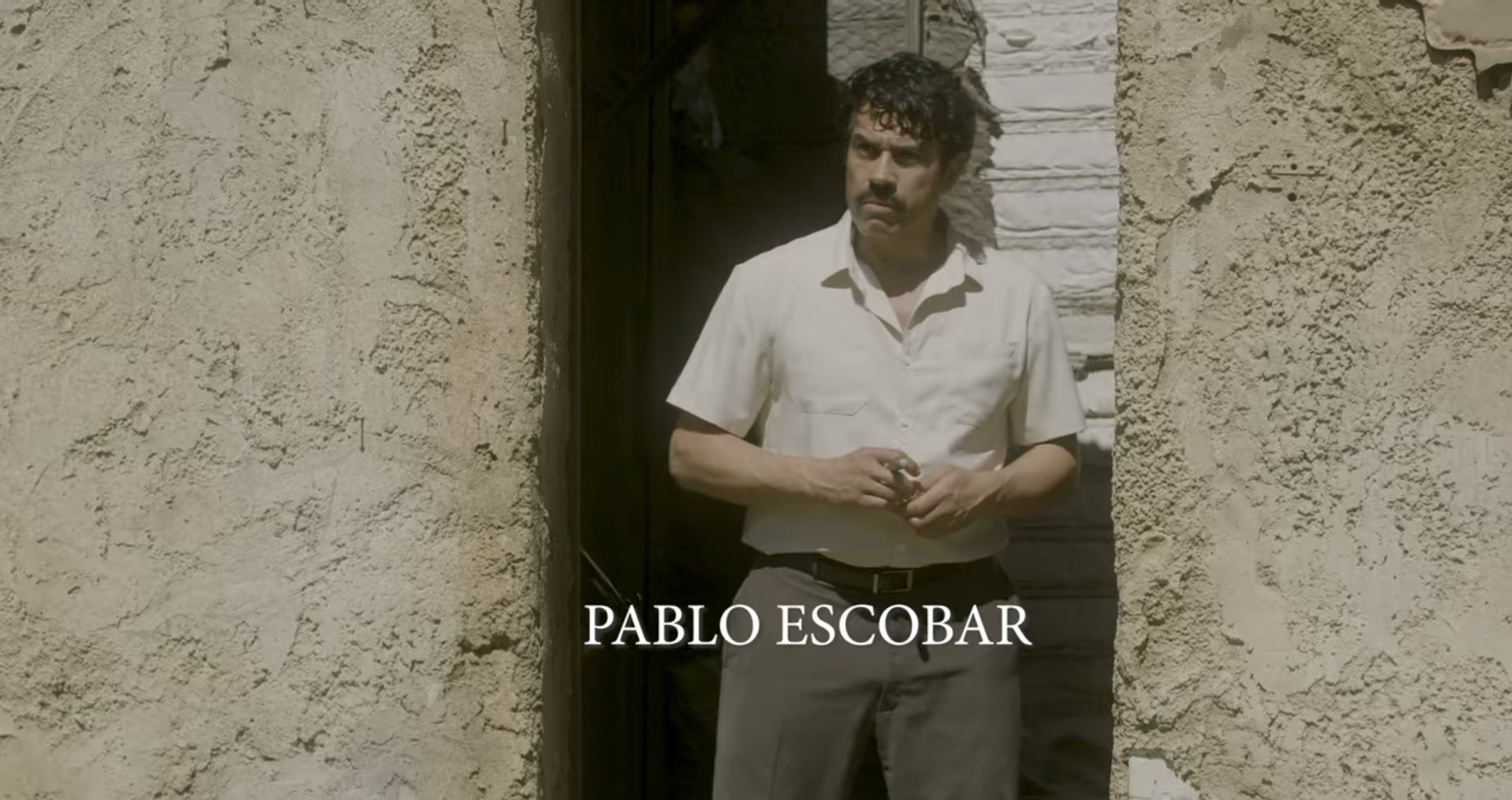 Man standing in doorway between two textured walls, wearing a white shirt, holding his hands together.