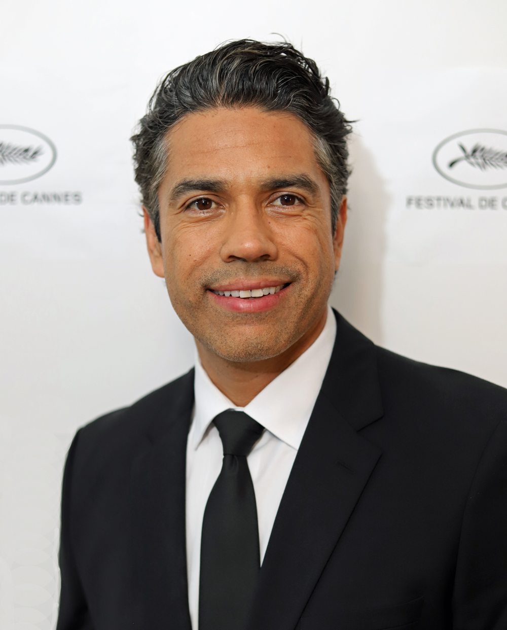 Man in a black suit and tie smiling, standing in front of a white background with logos.