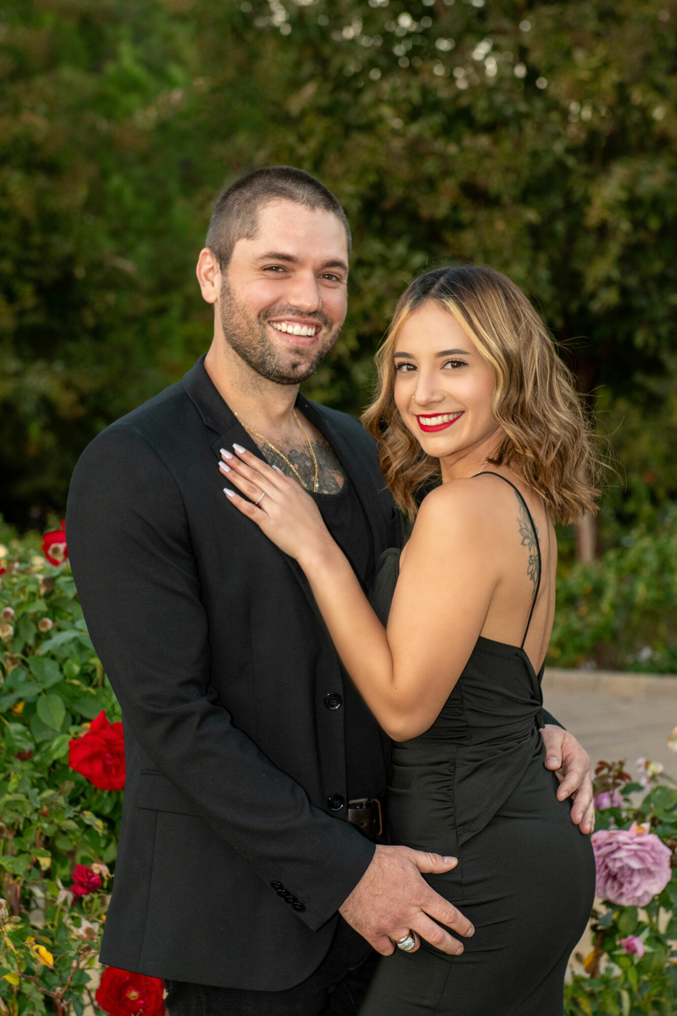 A smiling man and woman stand close together outdoors, surrounded by colorful flowers and greenery.