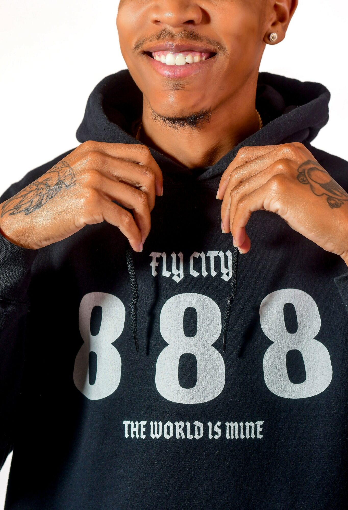 Person smiling, wearing a black hoodie with large gray numbers and text, holding the hoodie strings, tattoos visible on hands.