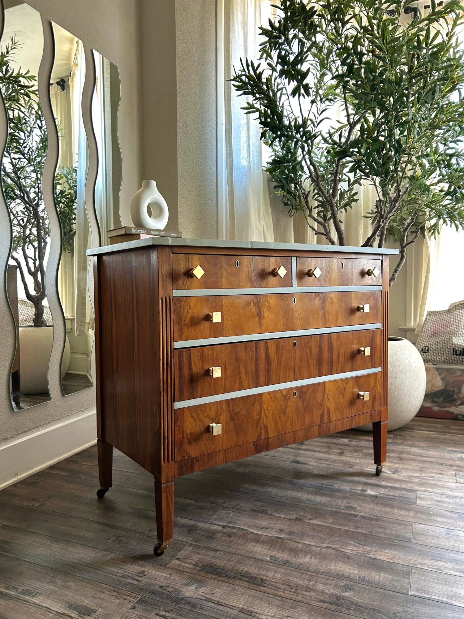Wooden dresser with four drawers and gold handles, positioned in a room with a large plant and decorative mirror.