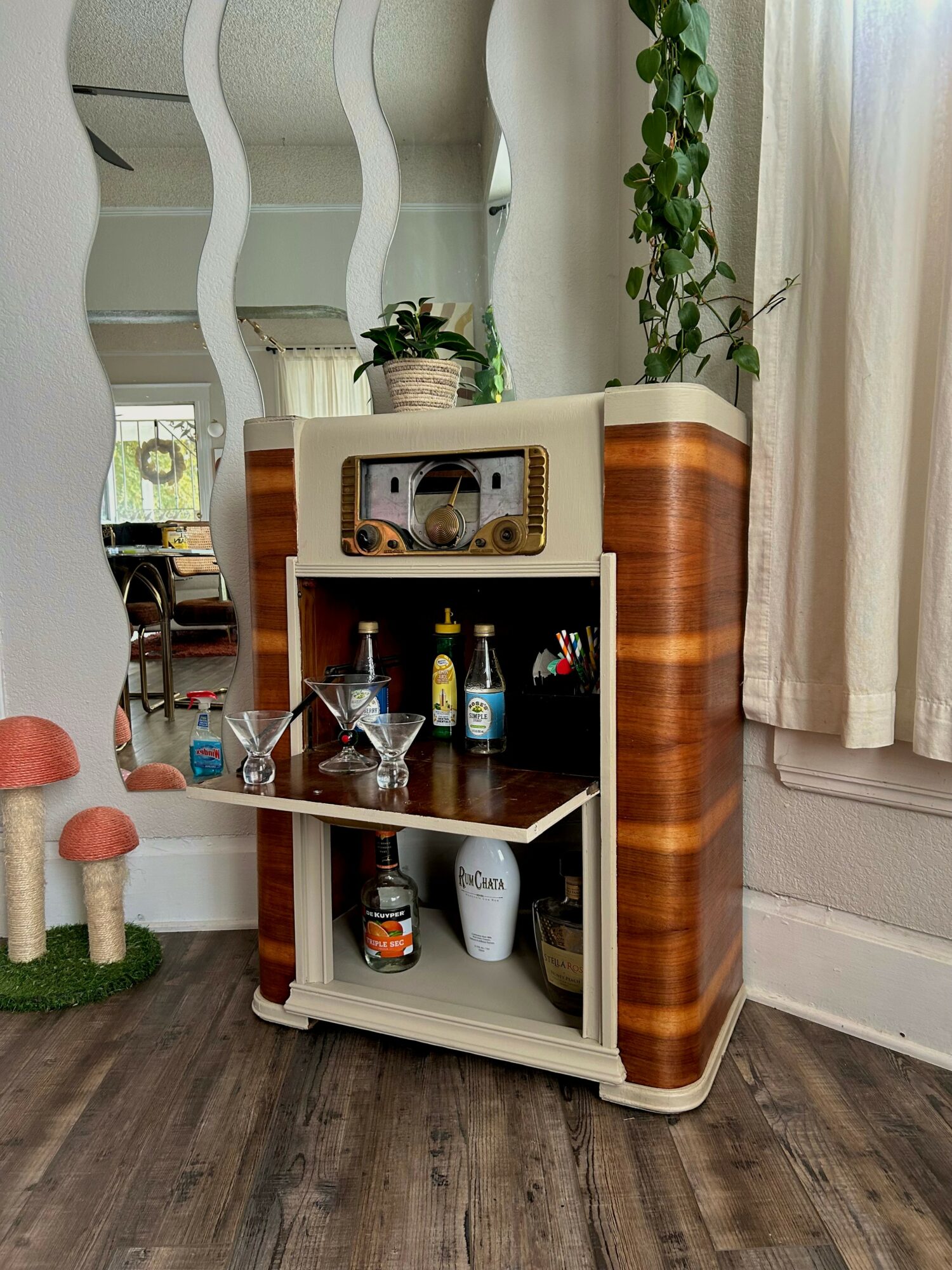 Wooden cabinet with shelves holding bottles, jars, and decorative items, next to a window with curtains and a mirror.
