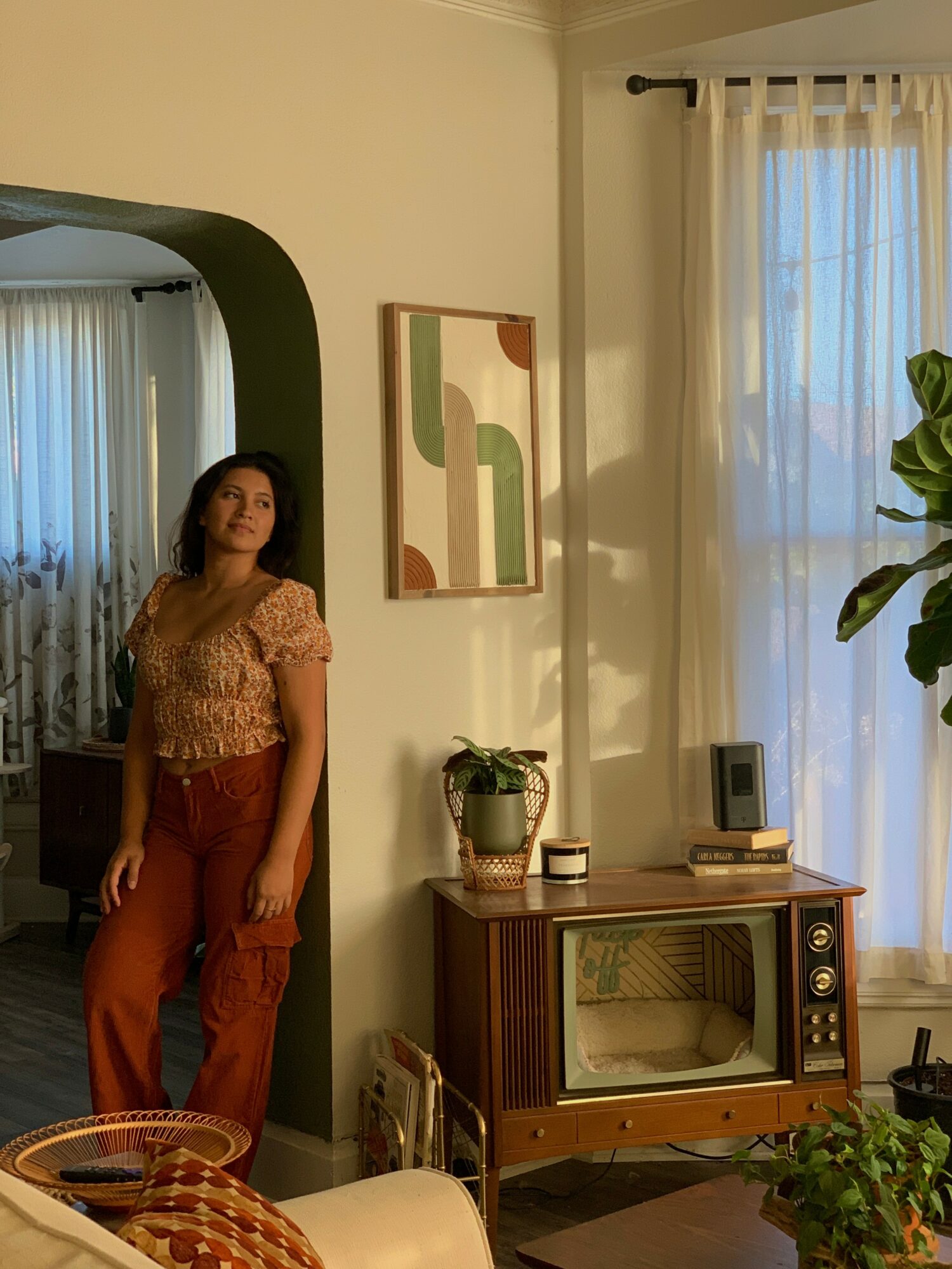 Woman standing near a wall with a framed abstract art piece, in a room with a window and a wooden cabinet.