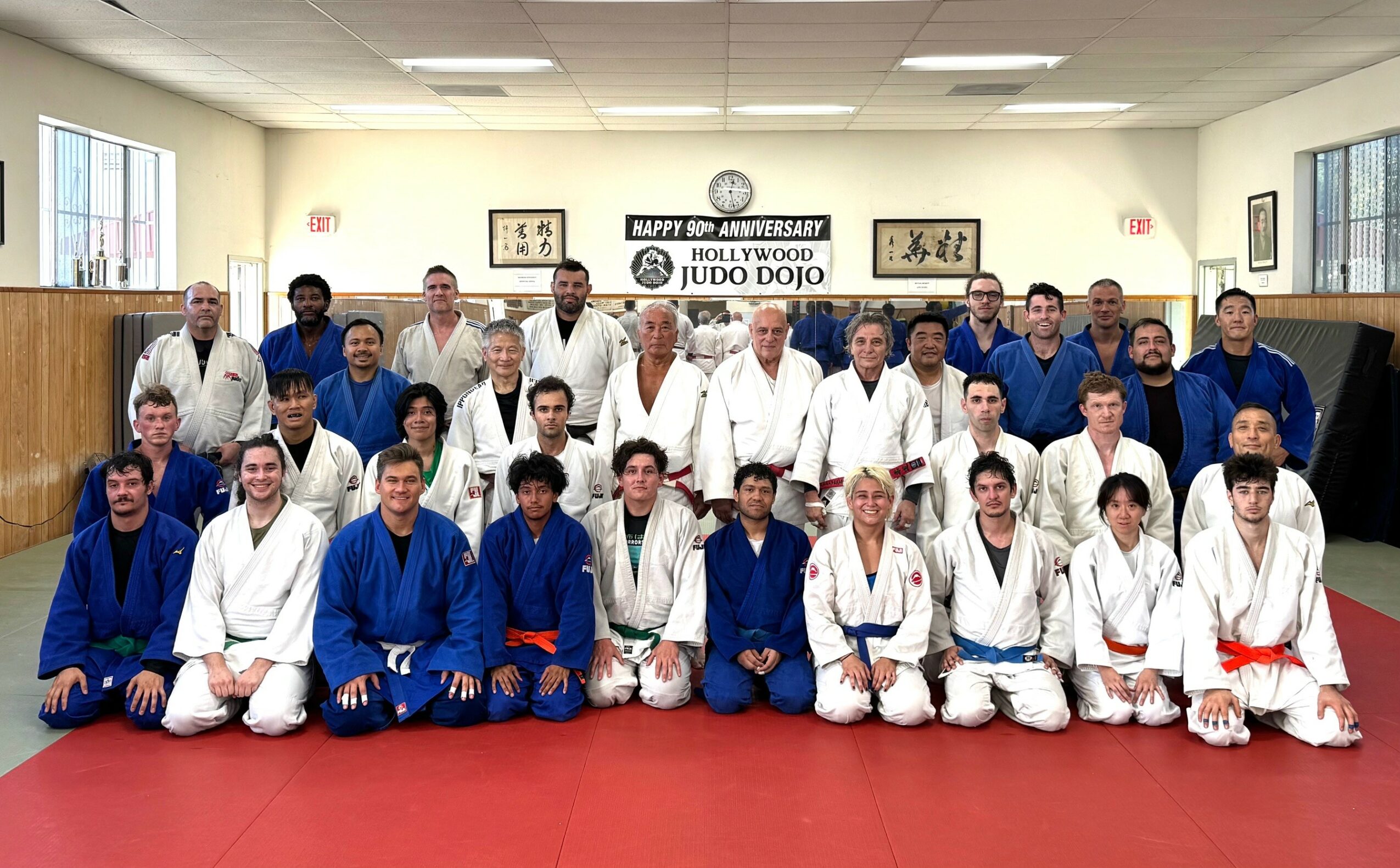 Group of people in judo uniforms and casual clothing in a dojo, with a banner and framed pictures on the wall behind.