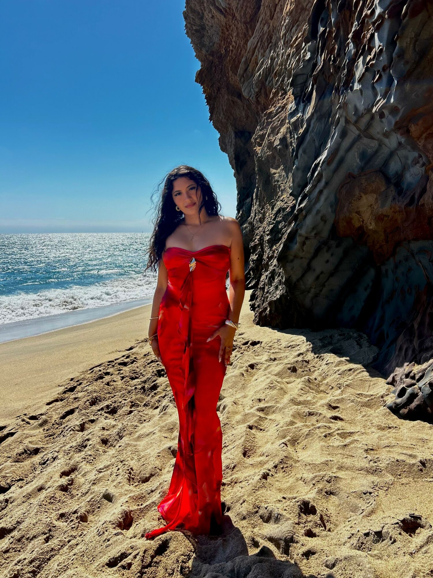Woman in red dress standing on sandy beach near large rock formation with ocean and blue sky in background.