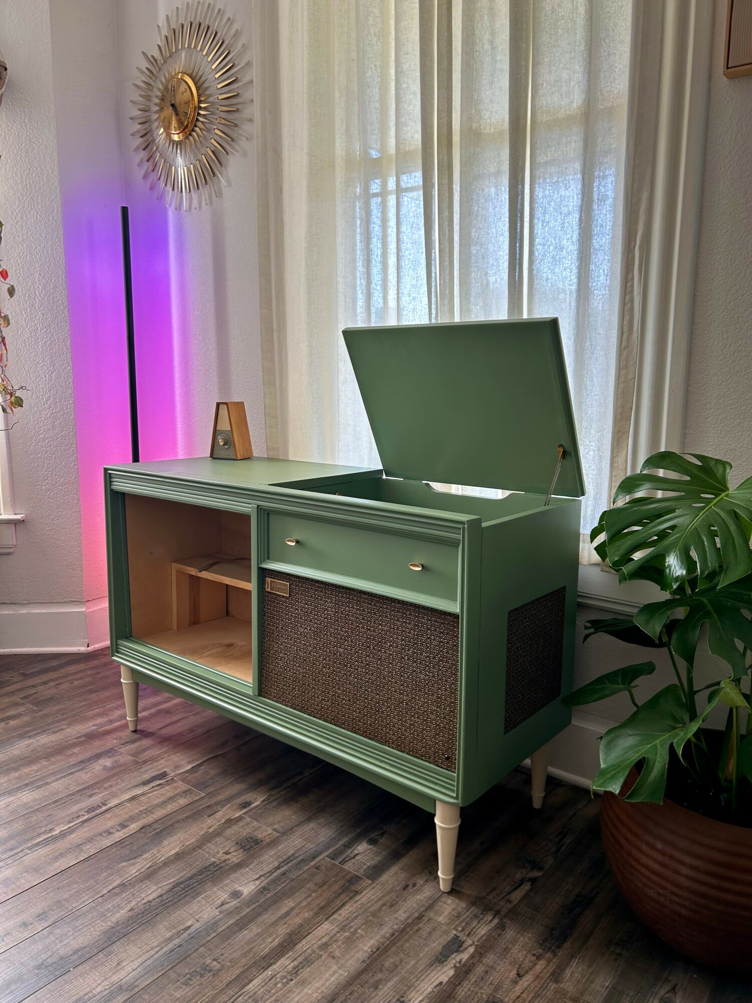 Green storage cabinet with open lid, drawers, and a large plant in a pot nearby, in a room with curtains and decorative wall art.