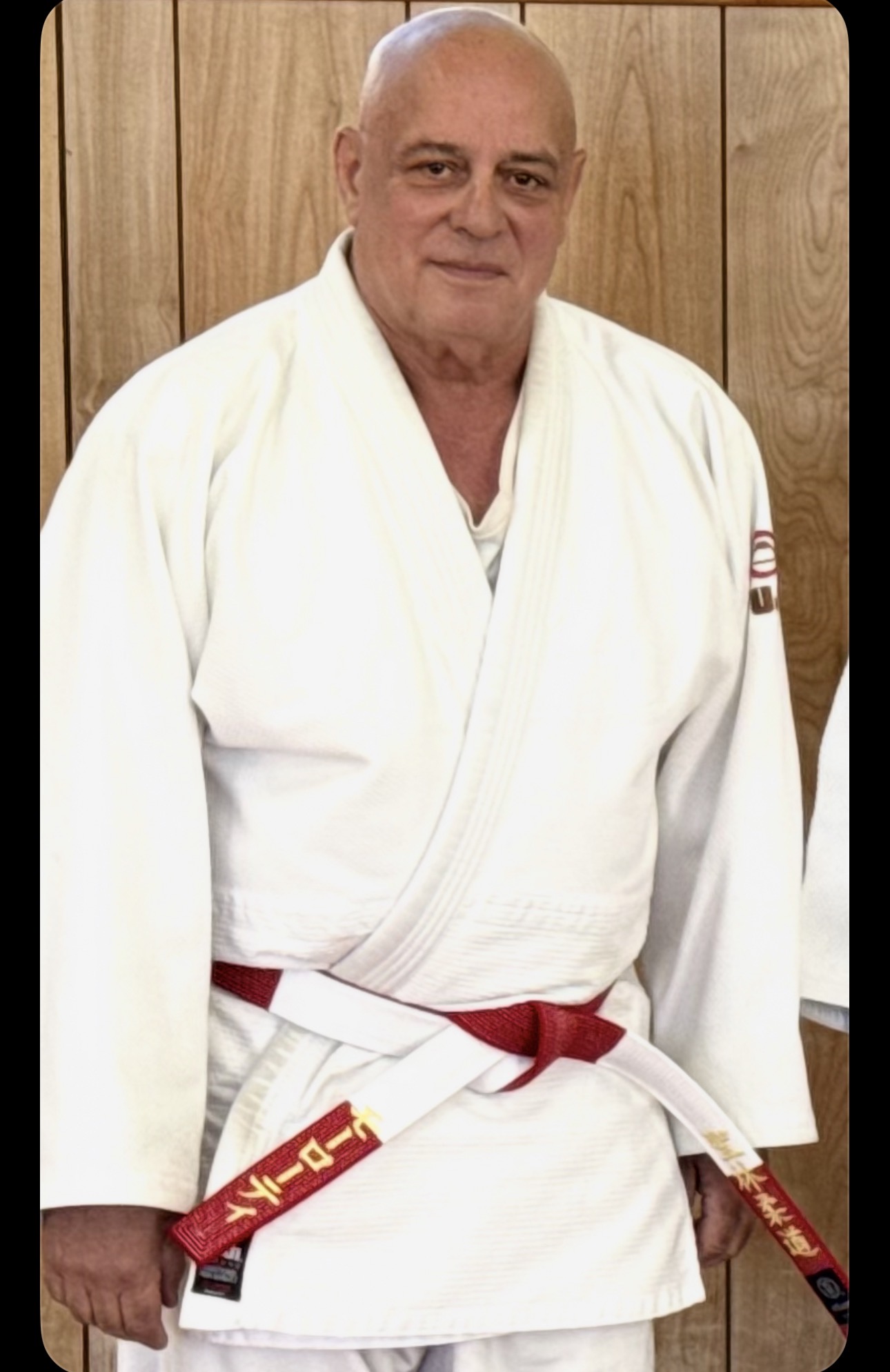 Man in a white martial arts uniform with a red belt, standing indoors against a wooden wall.