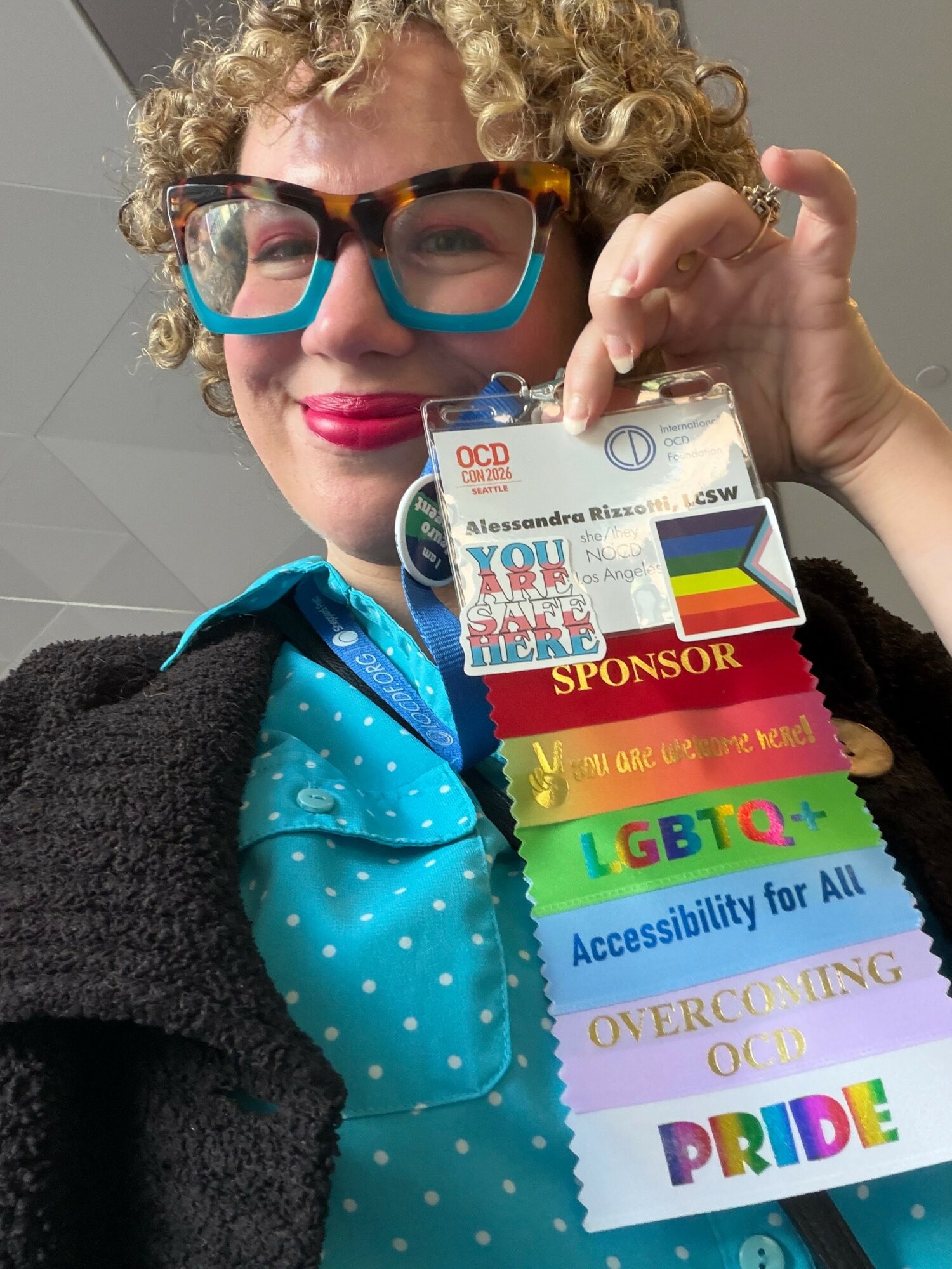 Person with curly hair and glasses holding a badge with rainbow-colored ribbons, smiling, wearing a blue polka-dot shirt and black jacket.