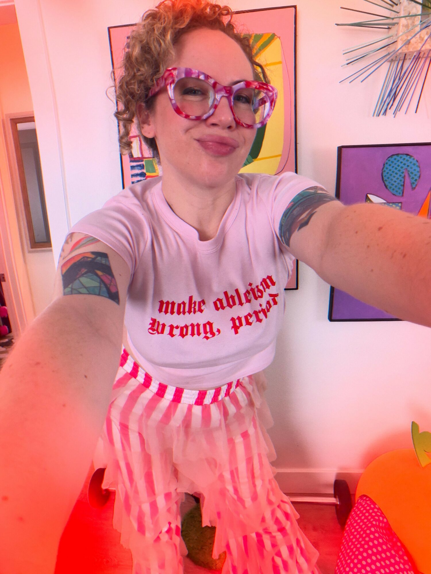 Person with curly hair wearing large pink glasses, a white t-shirt with red text, and a pink striped skirt, taking a selfie indoors.