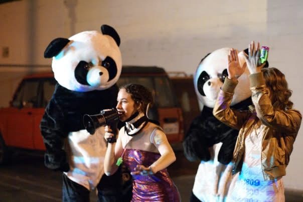 Two people in panda costumes and two women, one with a microphone, in an outdoor setting at night.