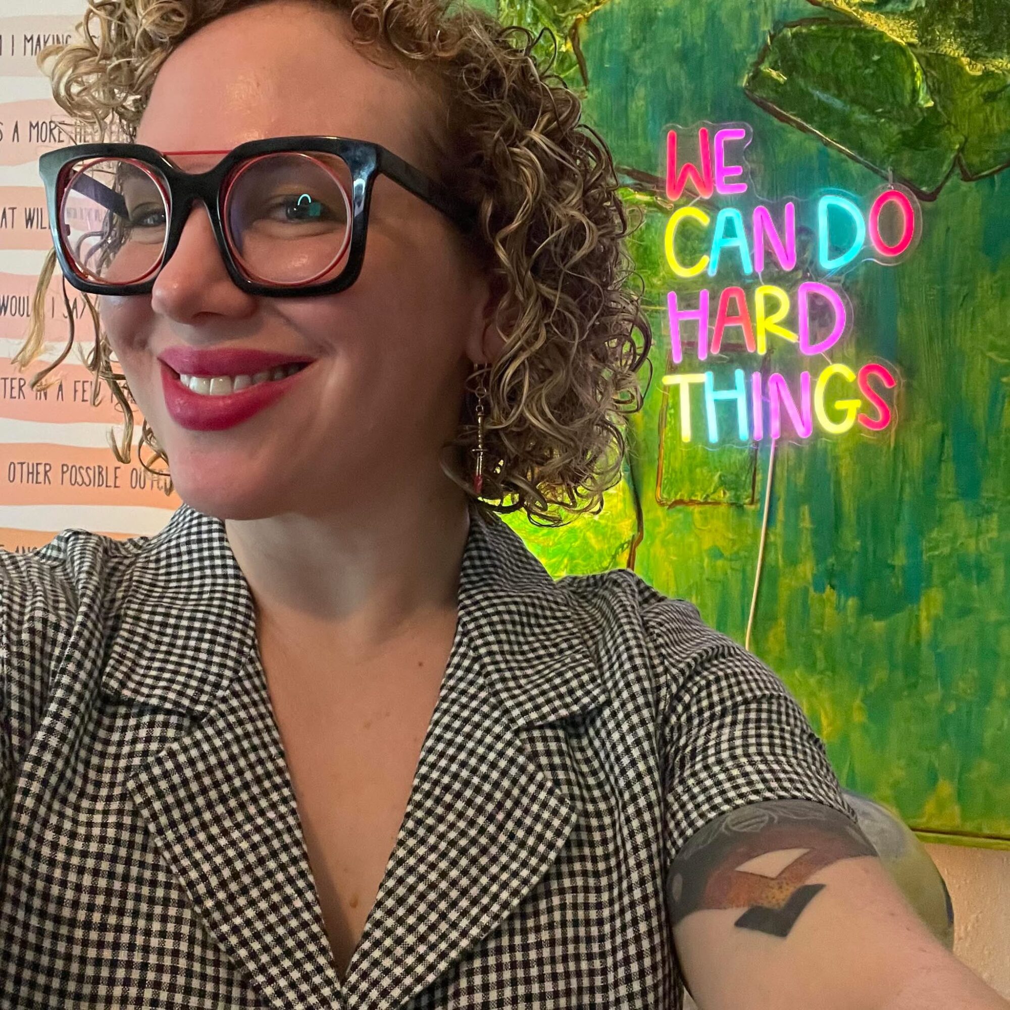 Woman with curly hair, glasses, and a checkered shirt smiling in front of a colorful background with neon text reading 'We Can Do Hard Things'.