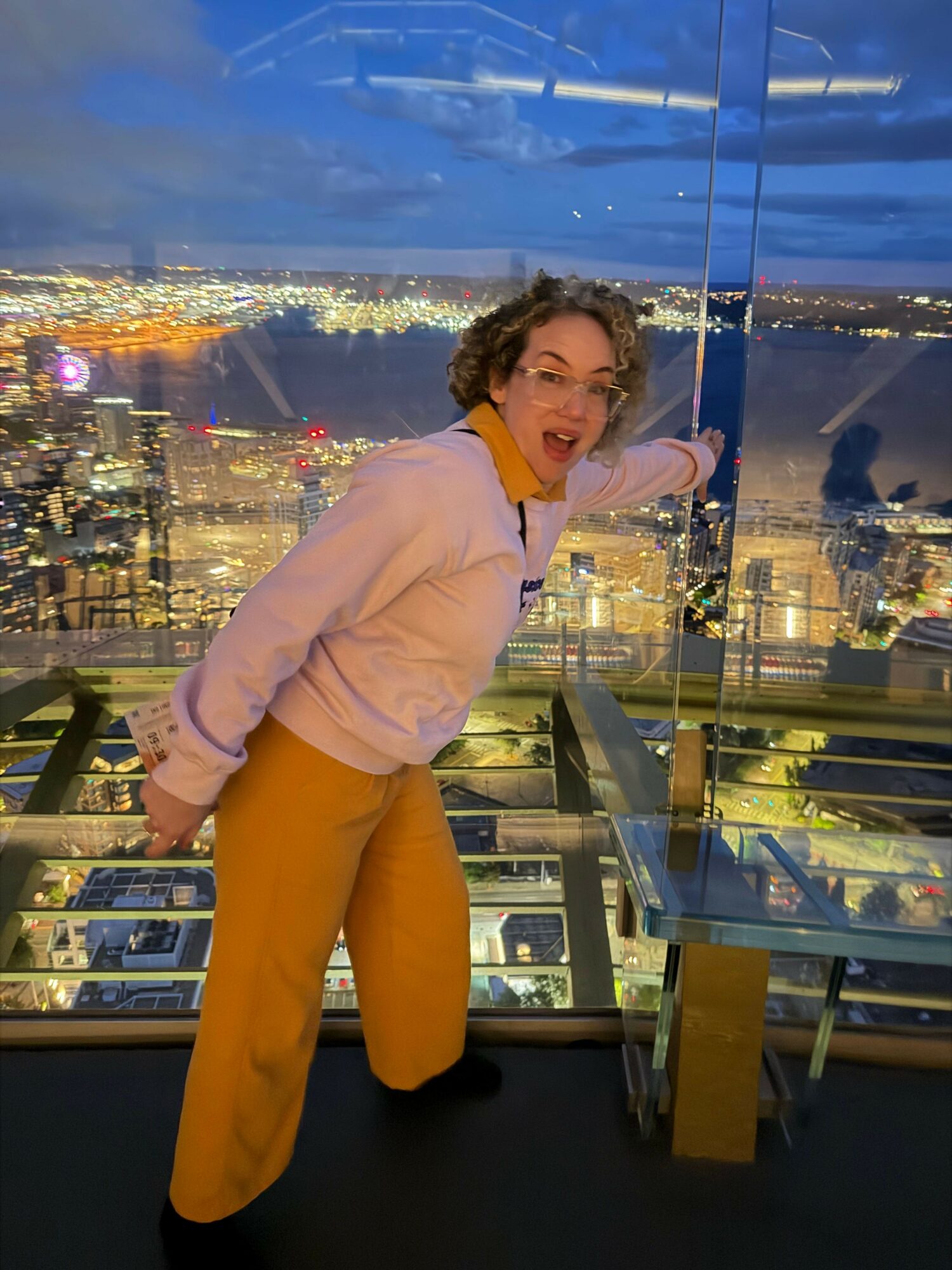 Person with curly hair in pink top and yellow pants standing in a display case, looking at the camera, with city lights in the background.