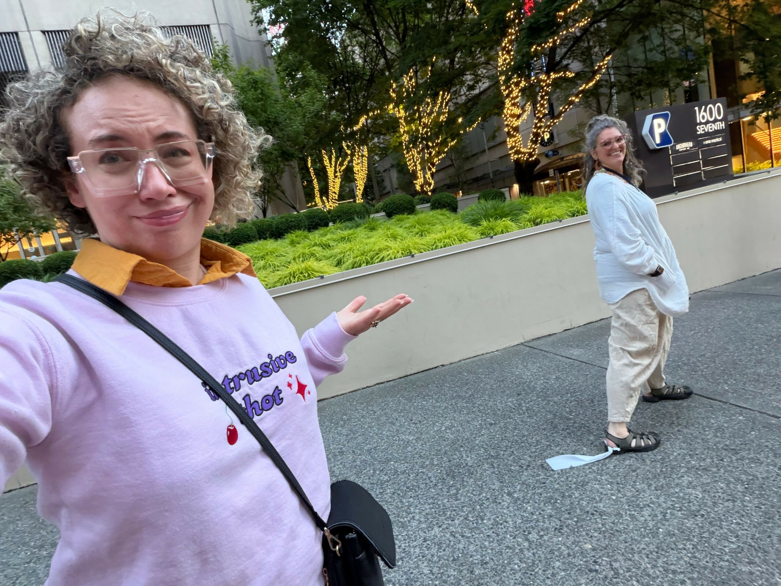 Woman taking selfie outdoors, smiling, pointing at a man standing on sidewalk, trees and greenery in background.