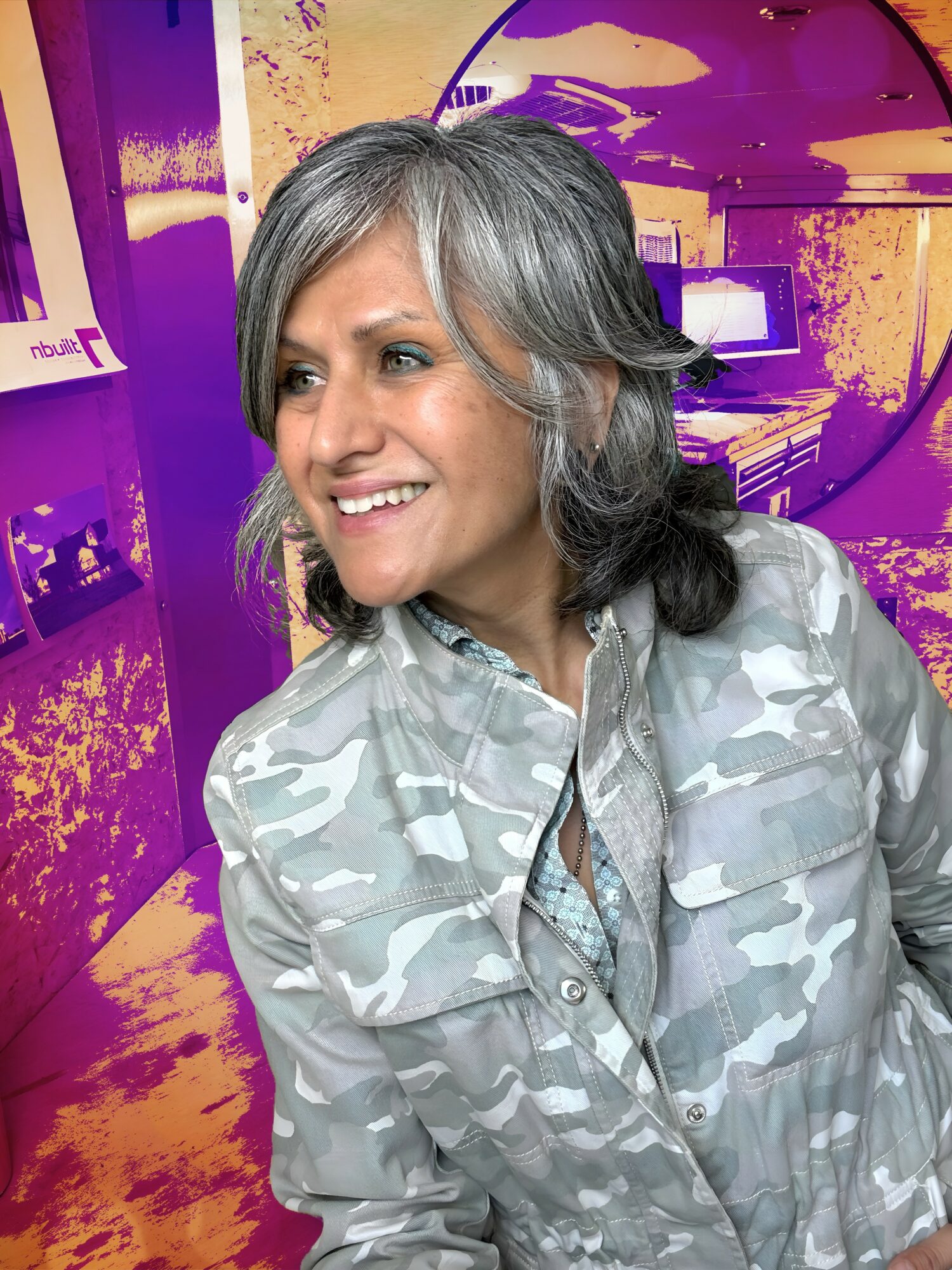 Smiling woman with gray hair in a camouflage shirt, standing in front of a colorful, abstract background.