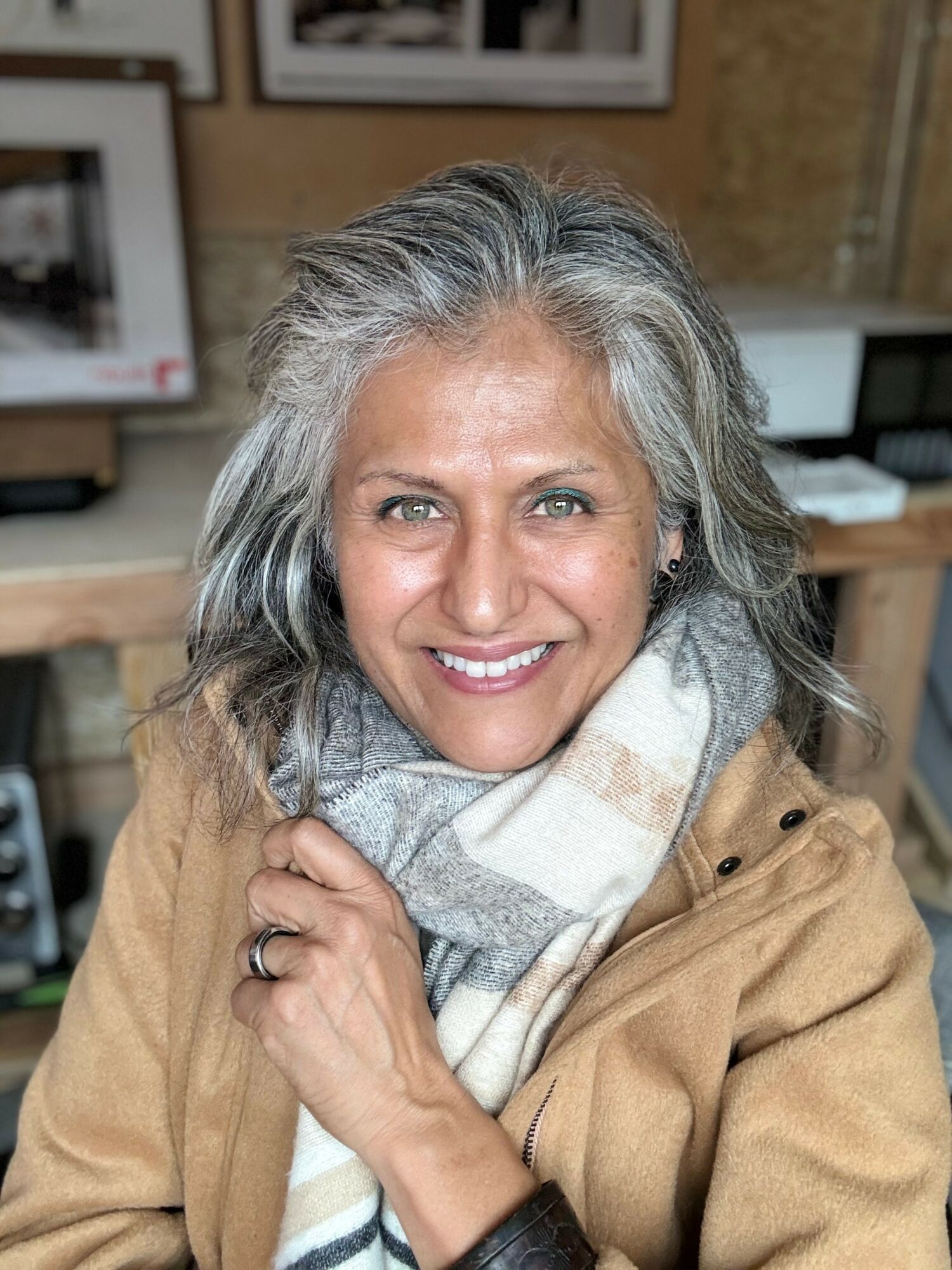 Smiling woman with gray hair wearing a beige coat and scarf, indoors with shelves and framed pictures behind her.