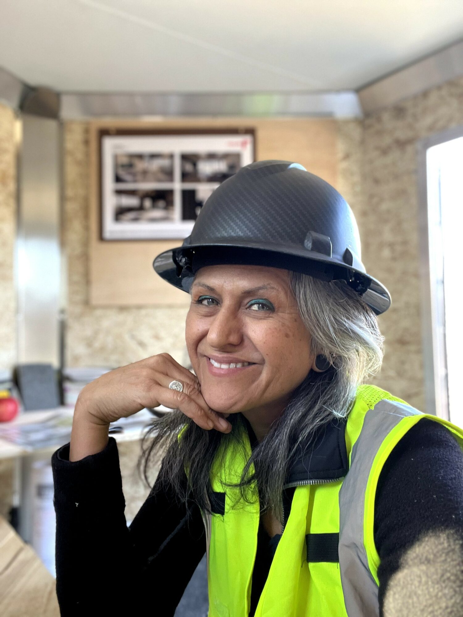 Woman wearing a black helmet and high-visibility vest, smiling indoors with a brick wall and window in background.